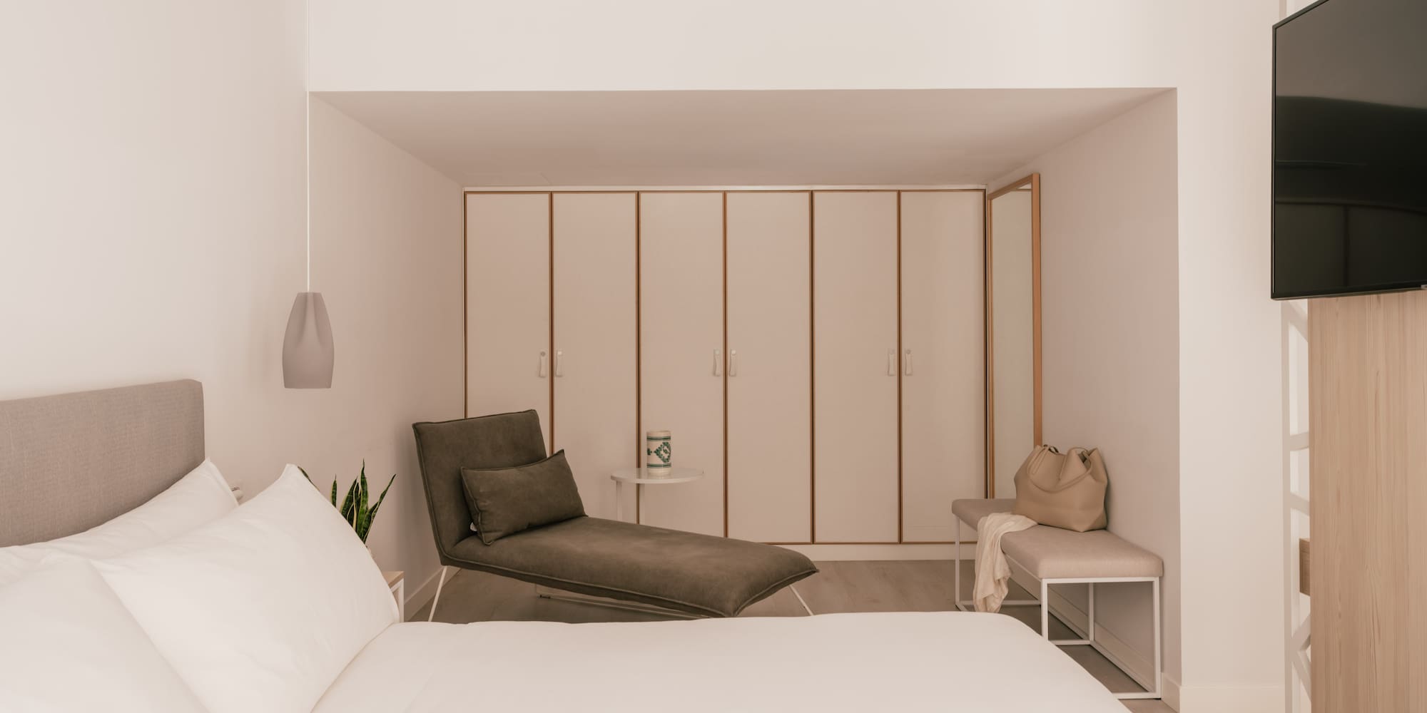 Modern hotel bedroom with white bed, grey chaise lounge, and built-in wardrobes.