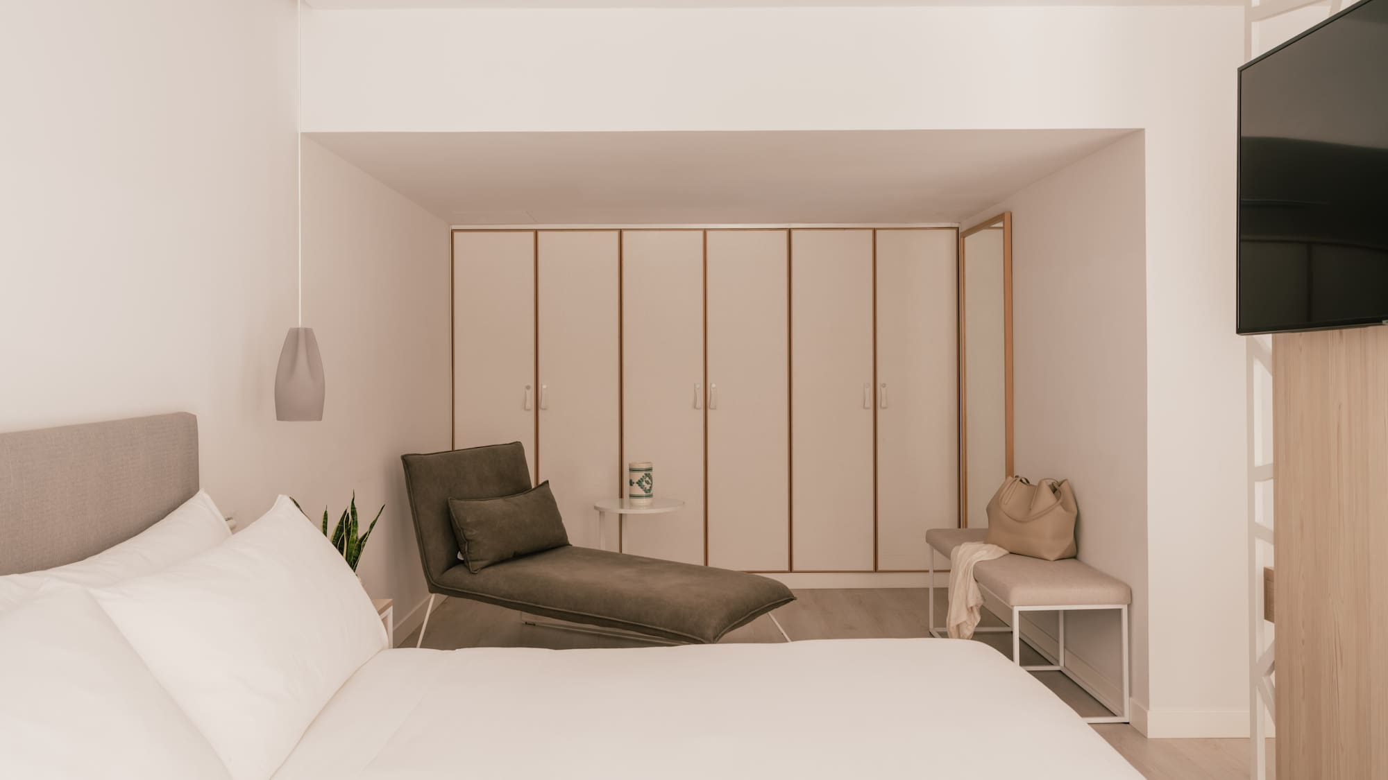 Modern hotel bedroom with white bed, grey chaise lounge, and built-in wardrobes.