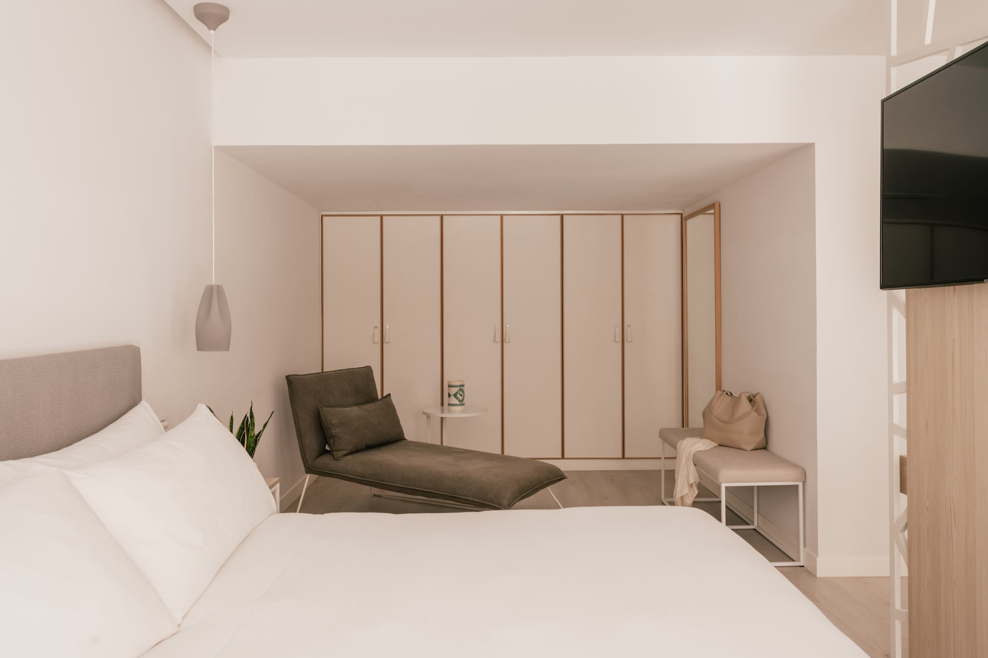 Modern hotel bedroom with white bed, grey chaise lounge, and built-in wardrobes.