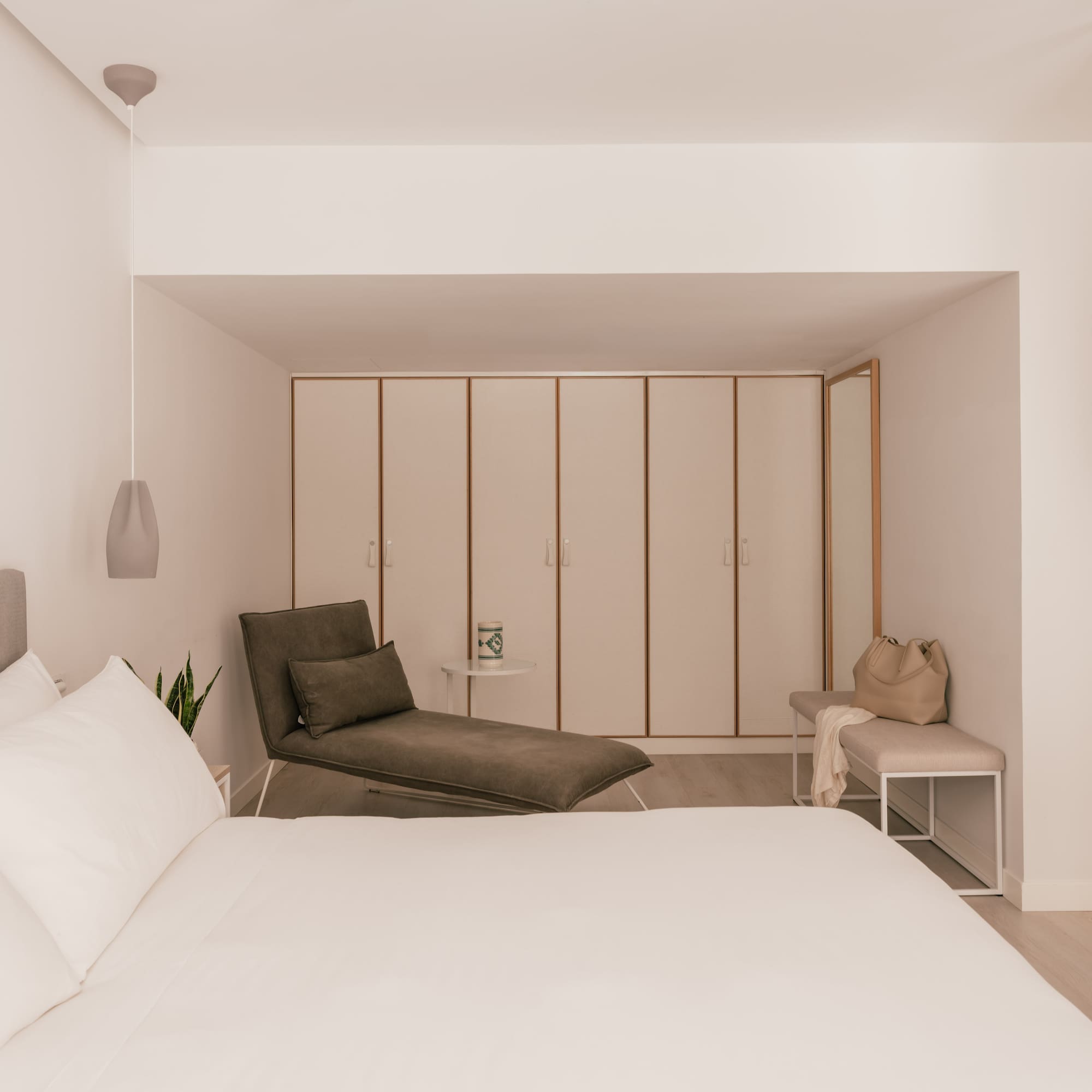 Modern hotel bedroom with white bed, grey chaise lounge, and built-in wardrobes.