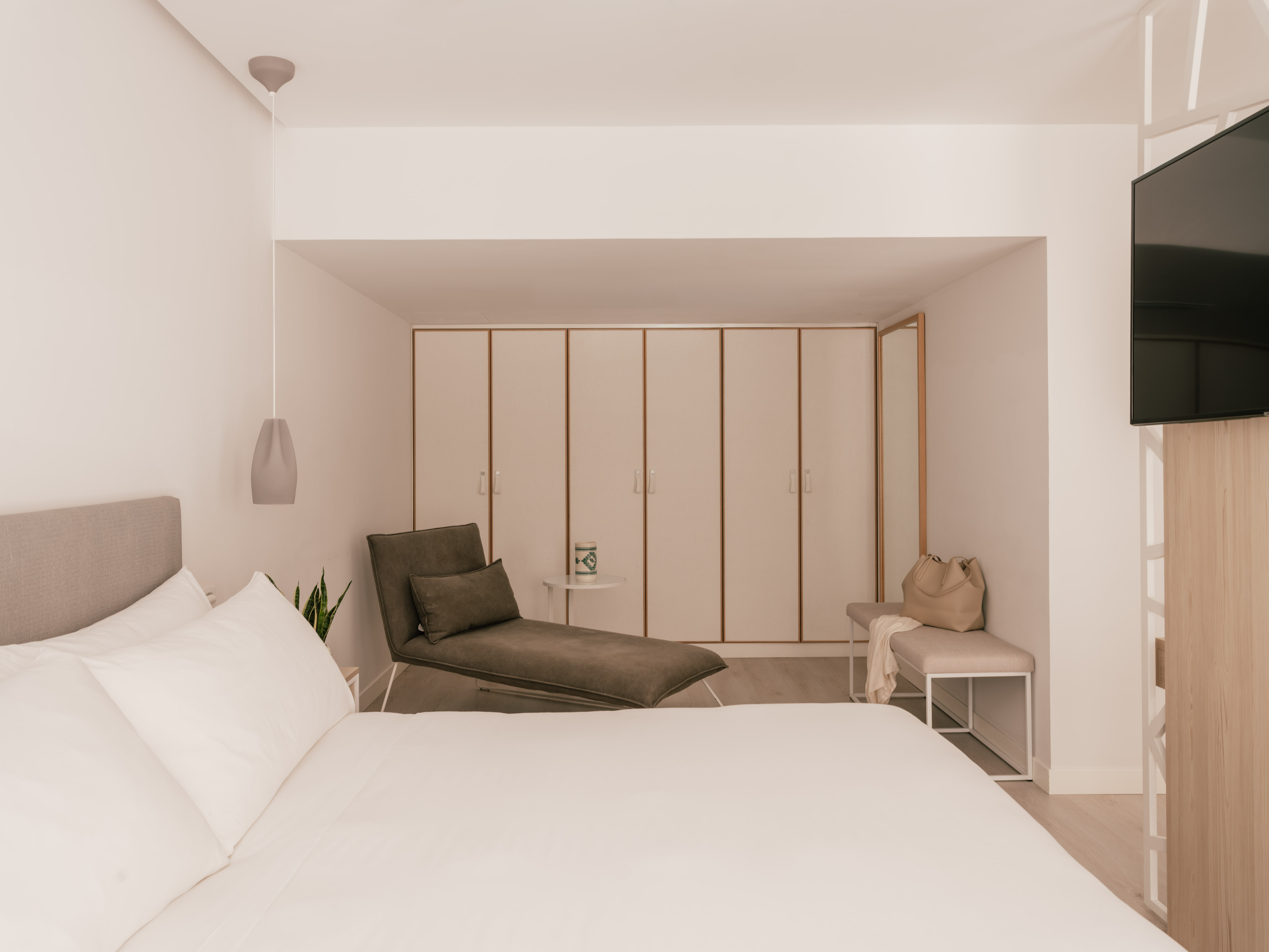 Modern hotel bedroom with white bed, grey chaise lounge, and built-in wardrobes.