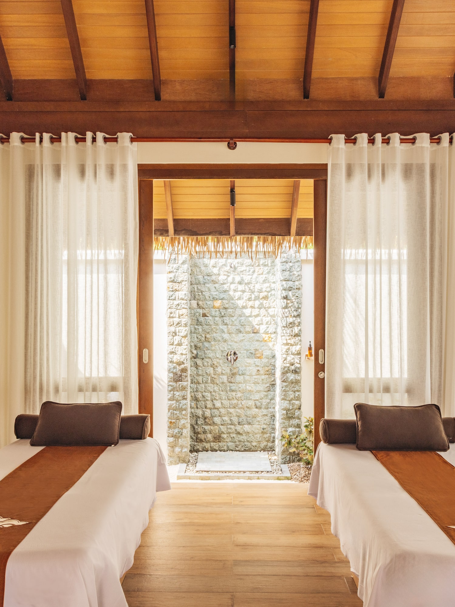 Tropical spa room with two massage tables, wood ceiling, and outdoor stone shower.