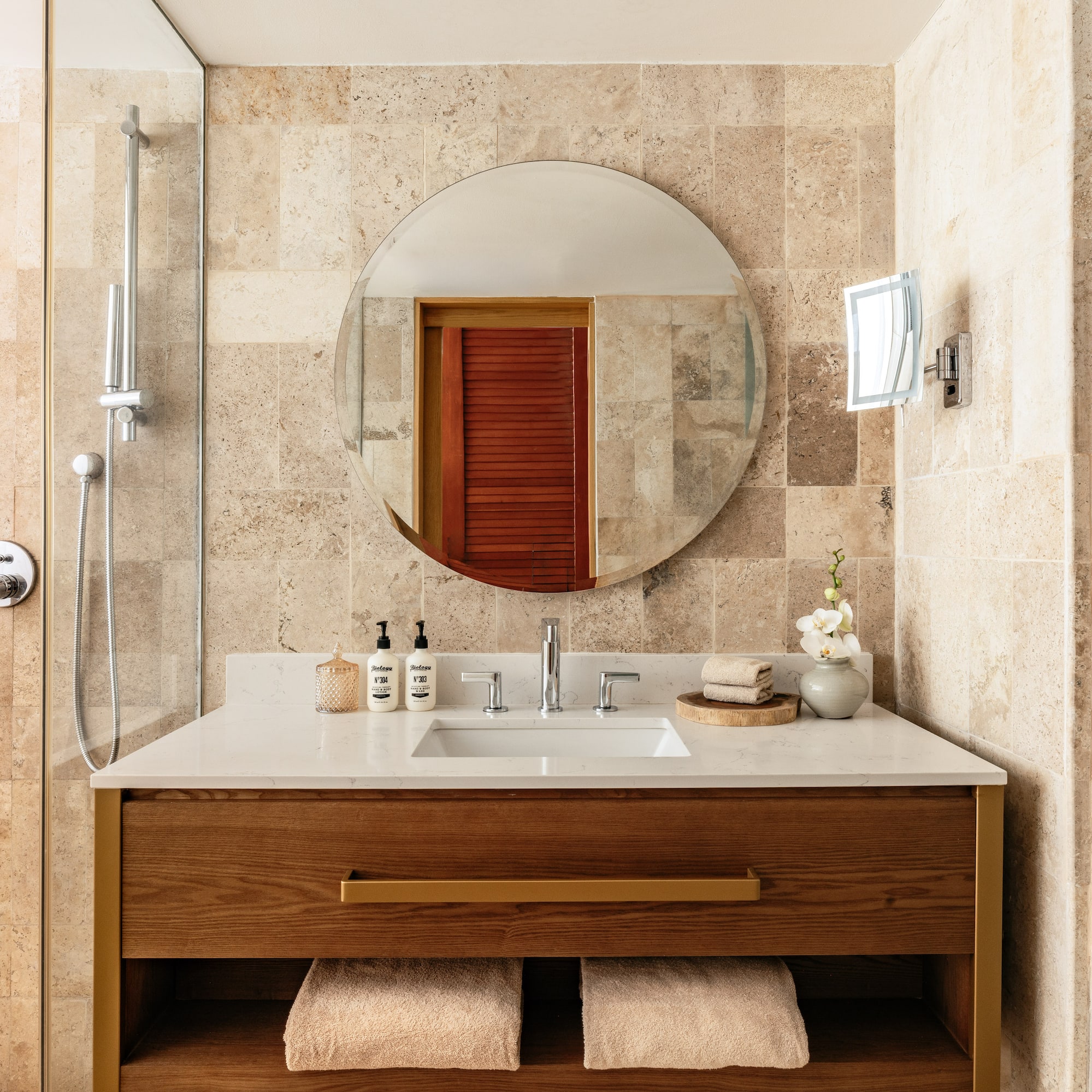 Elegant bathroom with travertine tiles, wood vanity, round mirror, and shower.