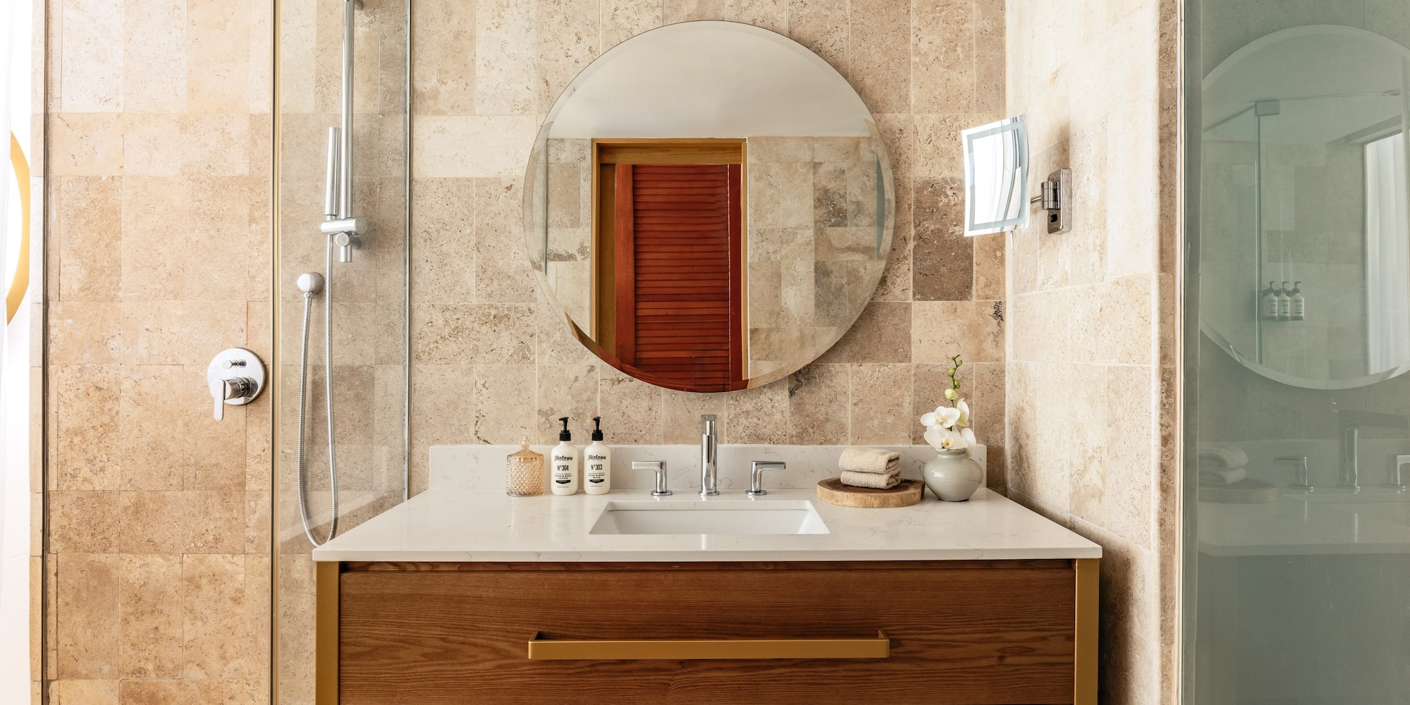 Elegant bathroom with travertine tiles, wood vanity, round mirror, and shower.