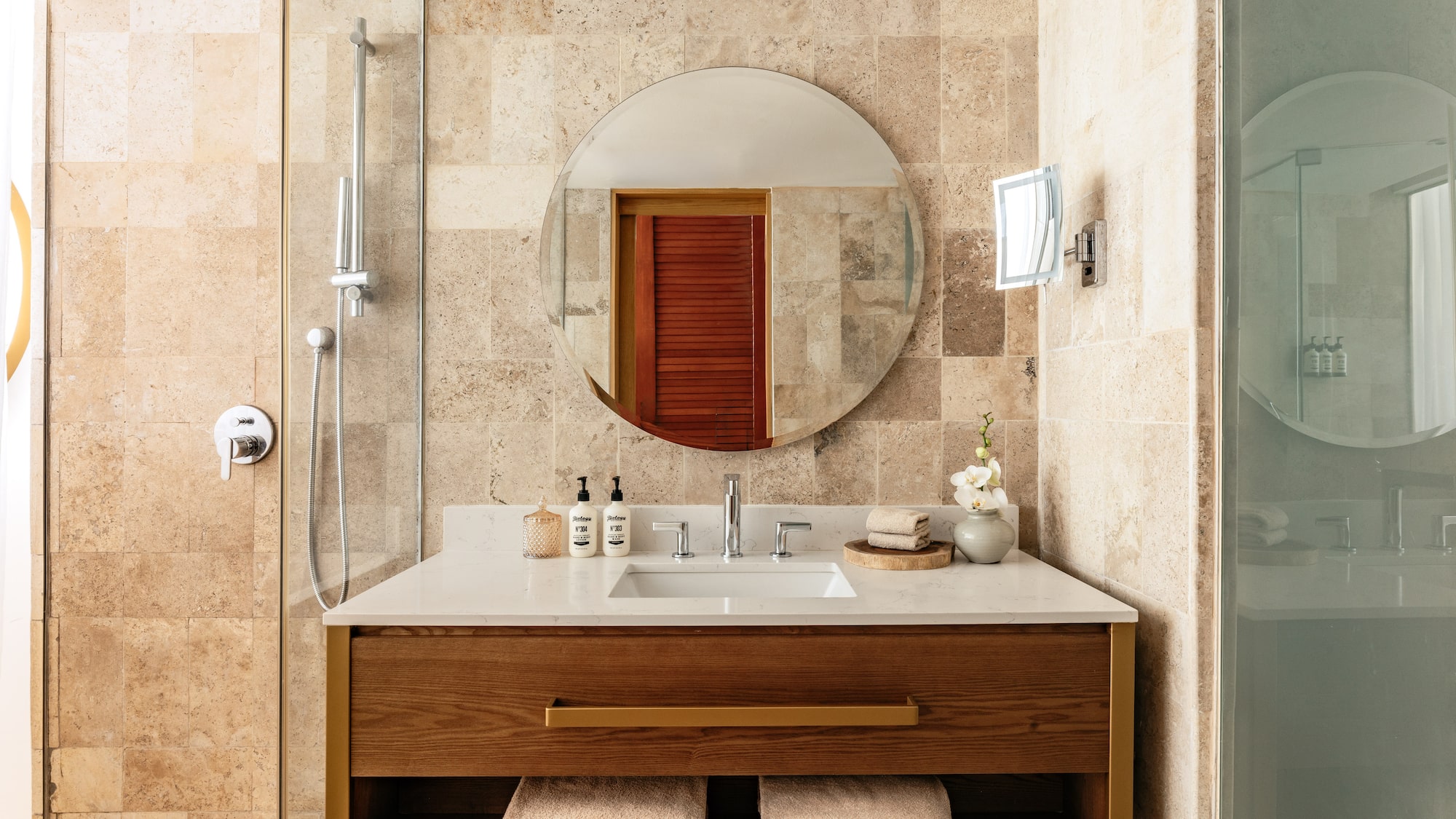 Elegant bathroom with travertine tiles, wood vanity, round mirror, and shower.