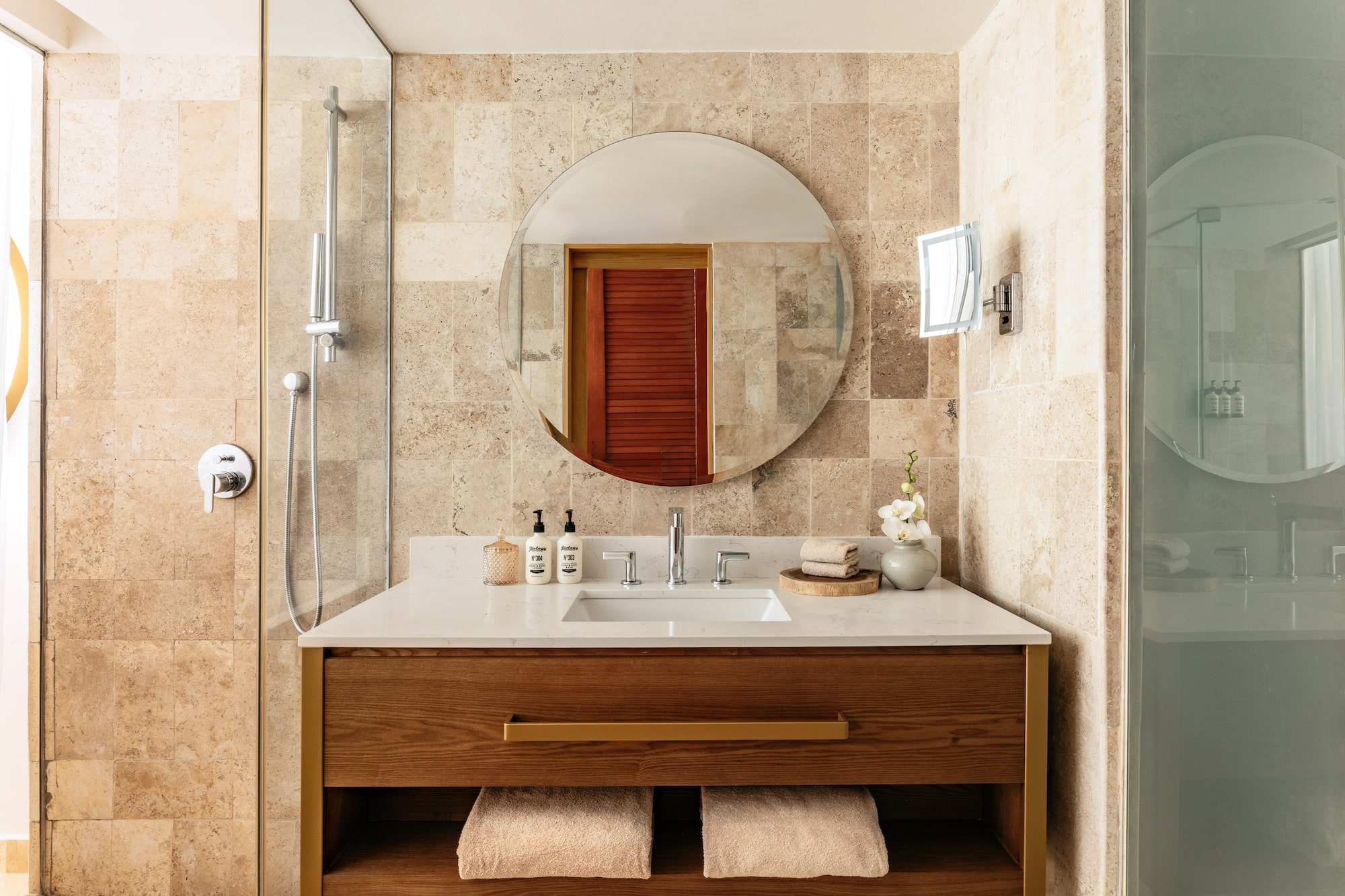 Elegant bathroom with travertine tiles, wood vanity, round mirror, and shower.