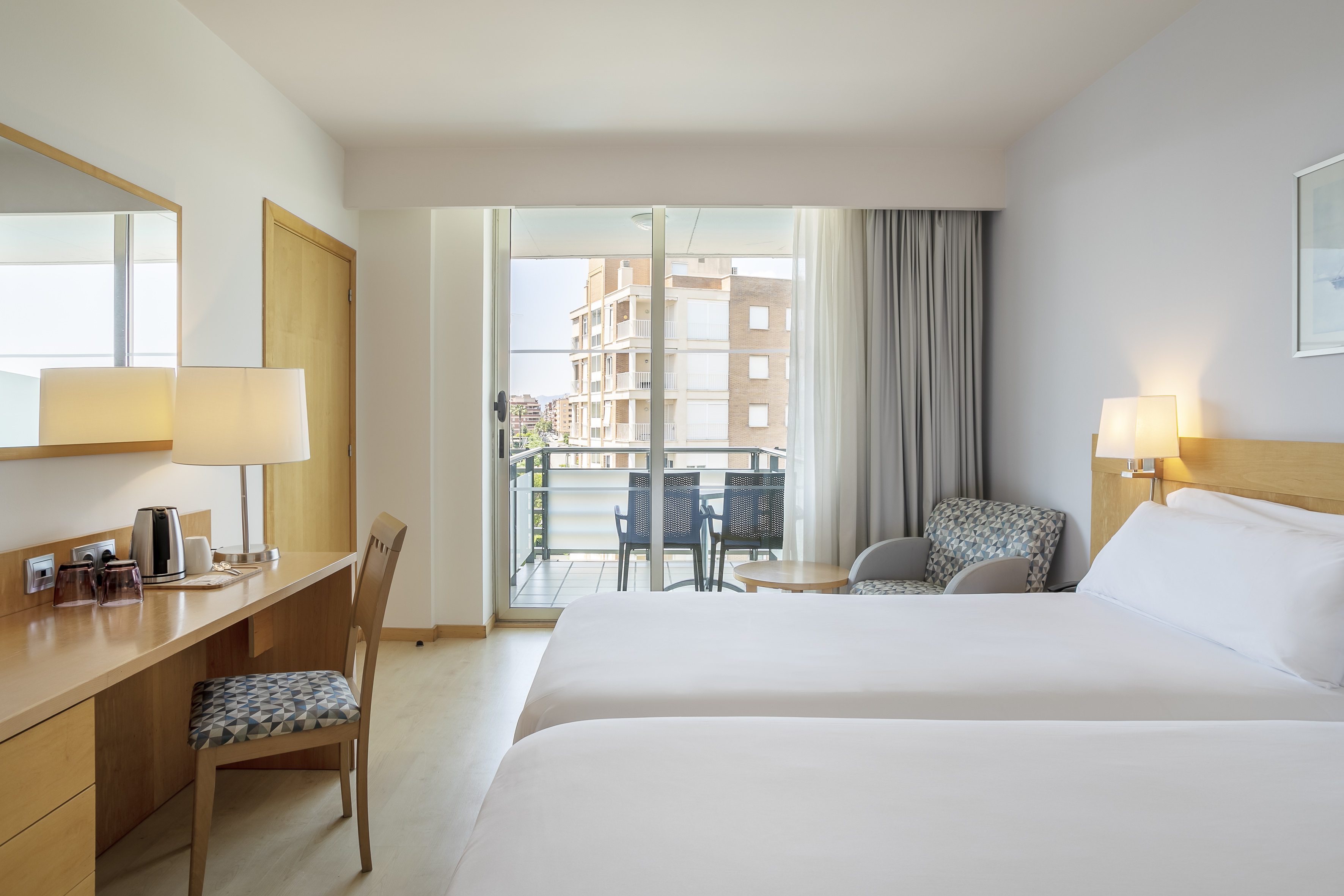 Bright hotel room with twin beds, desk, and balcony city view.