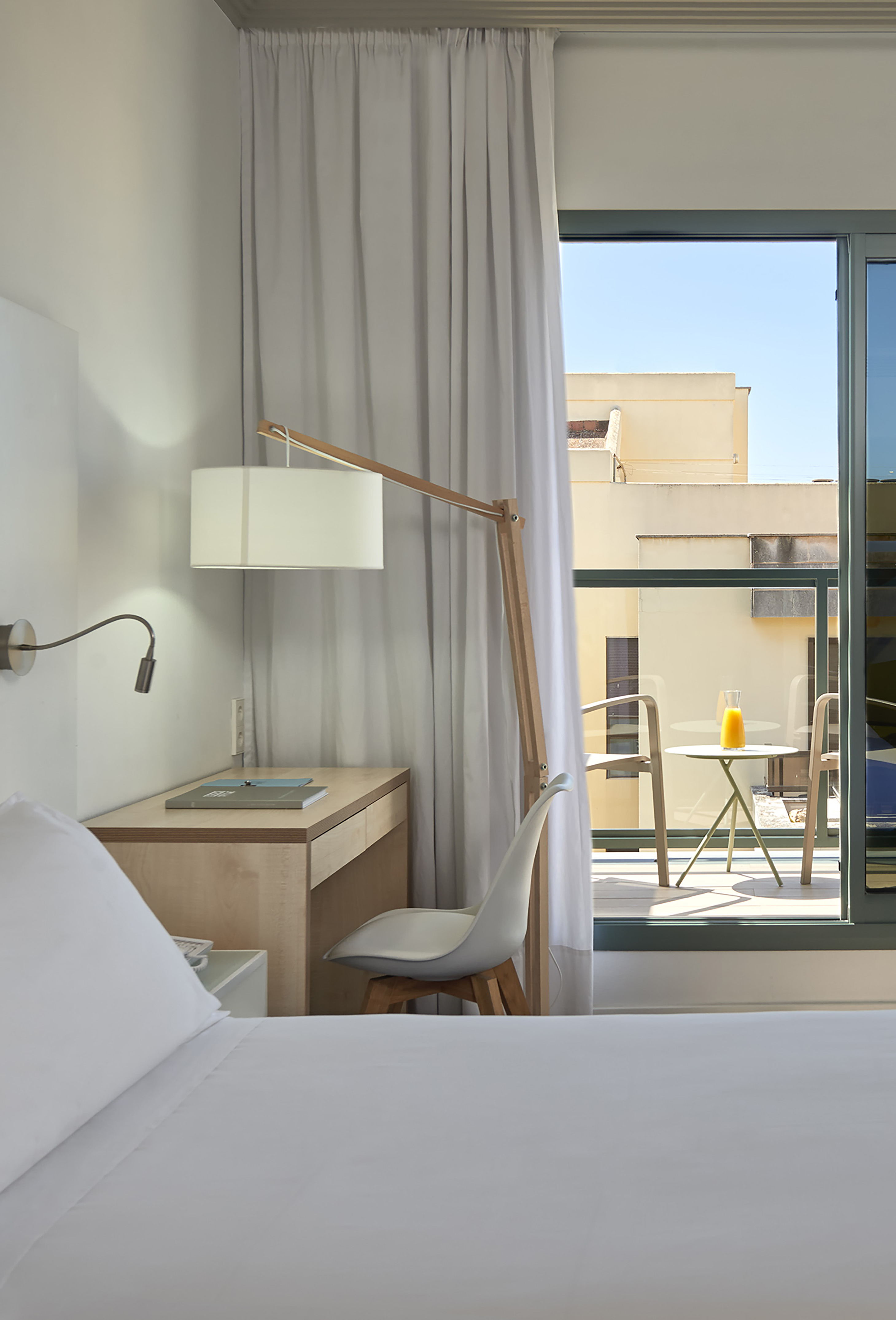 Bright modern hotel room with white bed, wood desk, and balcony with juice.