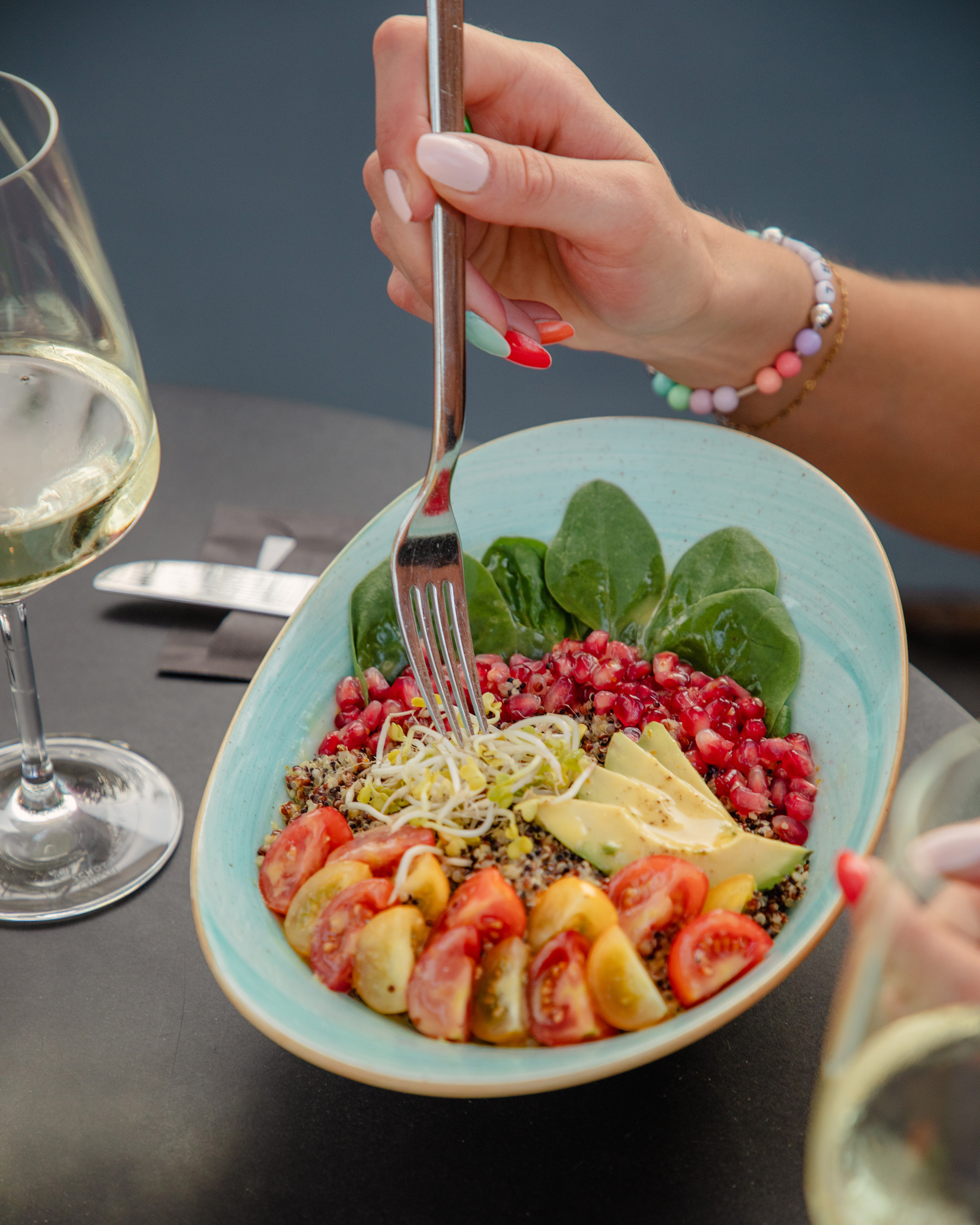 Fork in healthy quinoa bowl: avocado, tomatoes, spinach, pomegranate, wine.