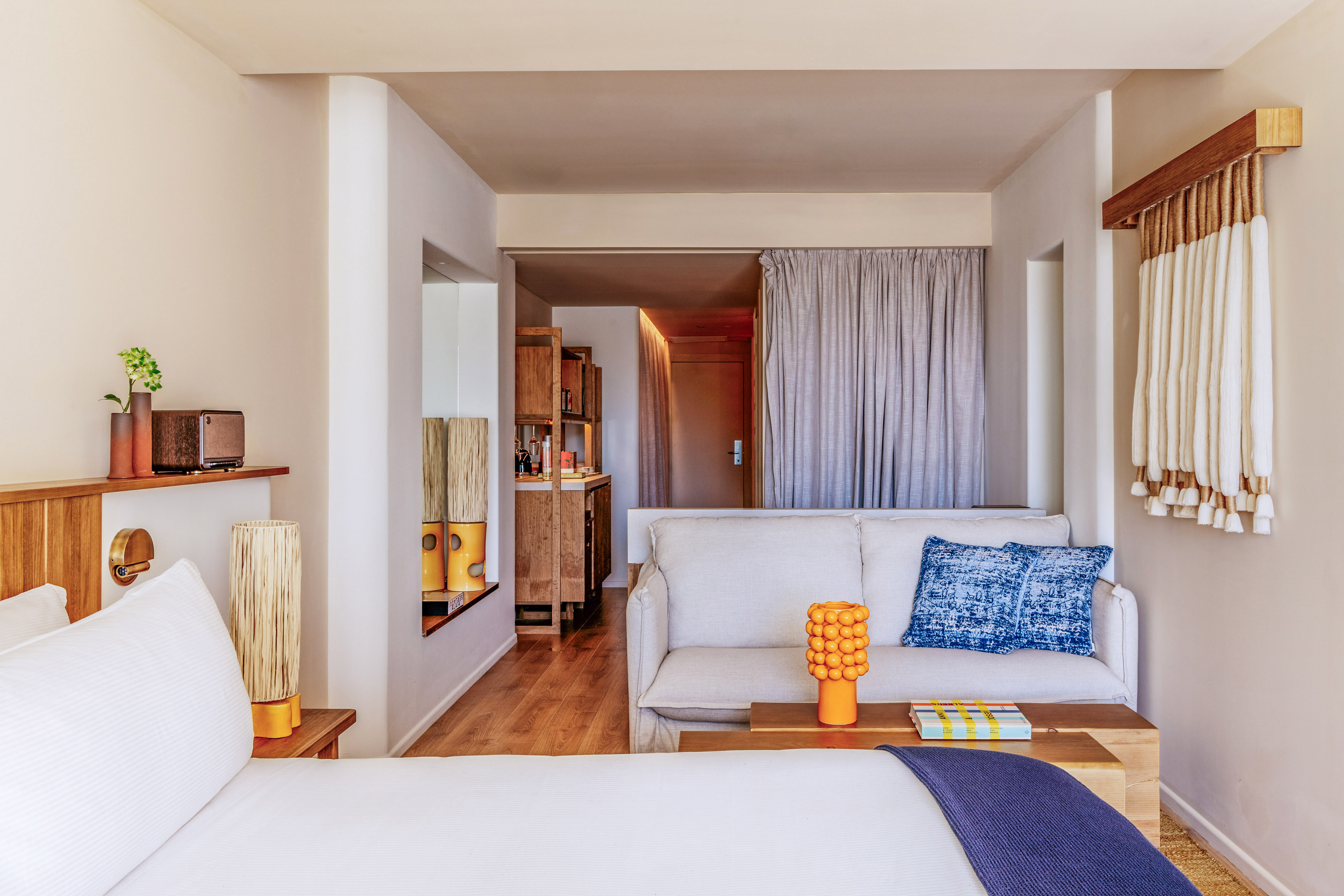 Modern hotel studio room with bed, sofa, and wood kitchenette in neutral tones.