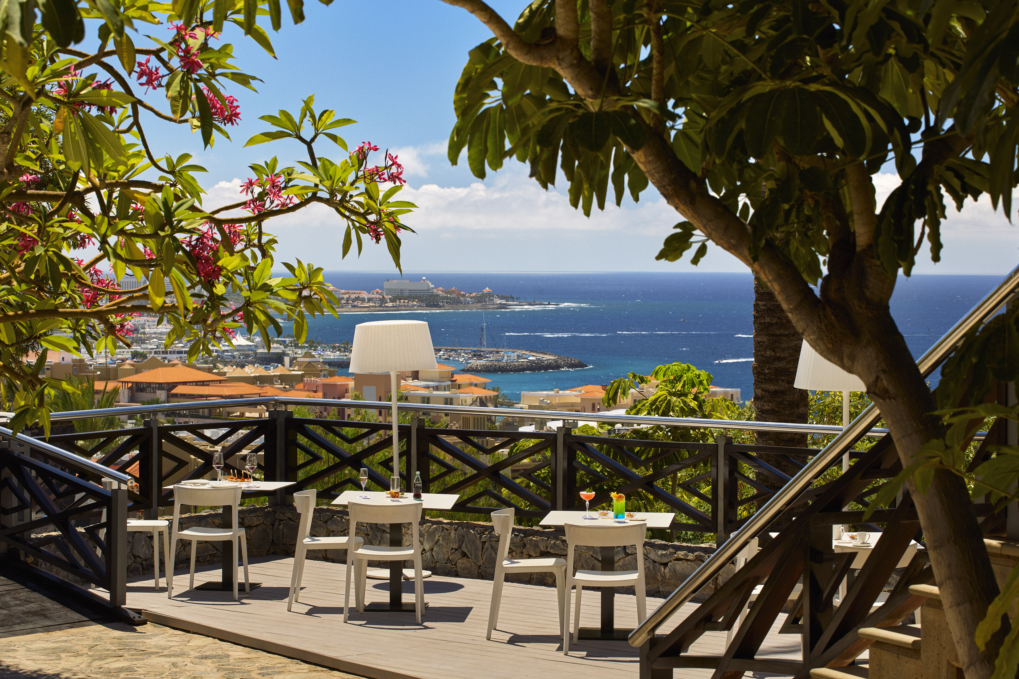 Coastal resort terrace with ocean view, dining tables, and tropical flowers.