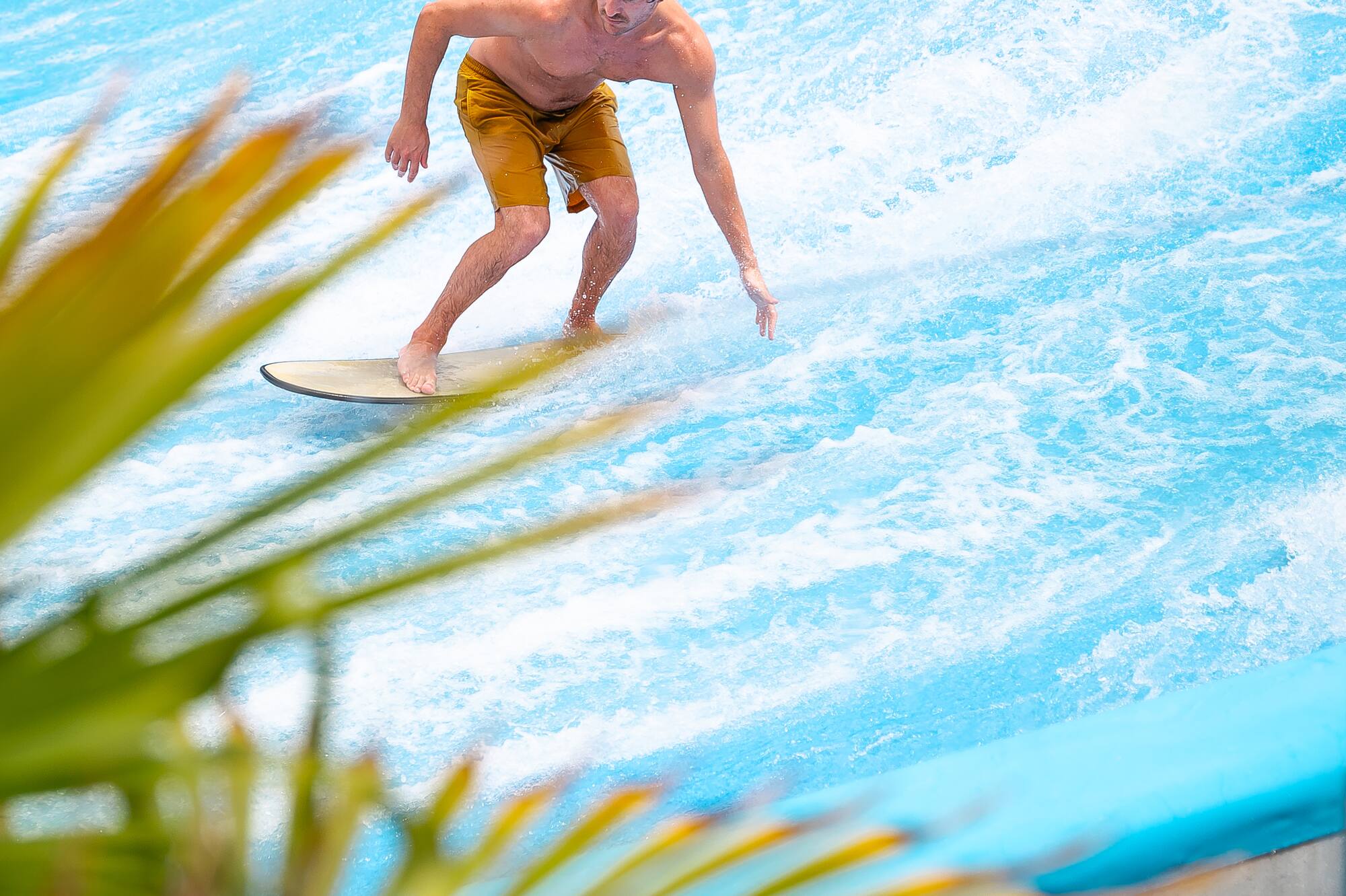 Young man surfing on a bright blue flowrider simulator with palm leaves.
