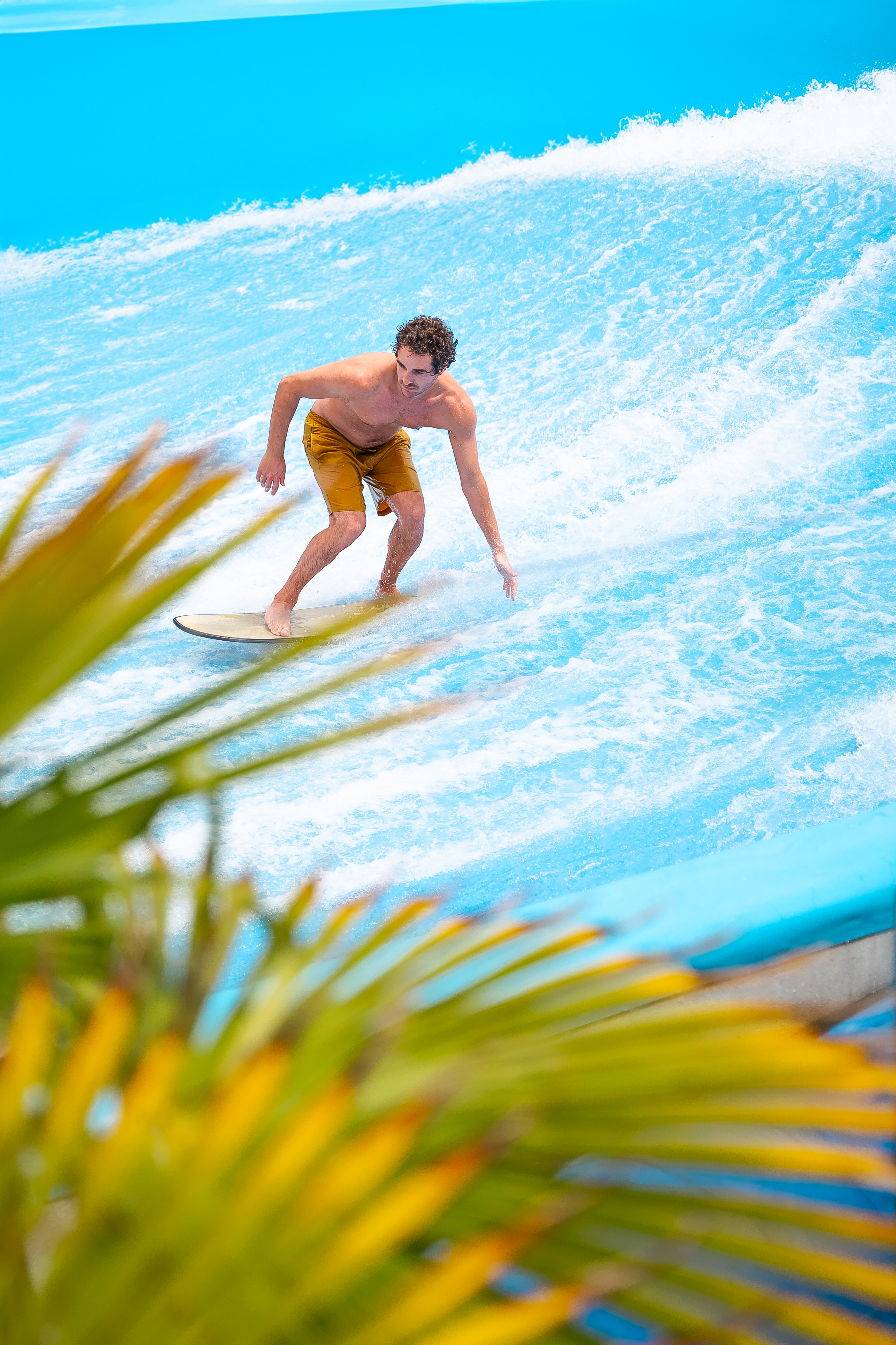 Young man surfing on a bright blue flowrider simulator with palm leaves.