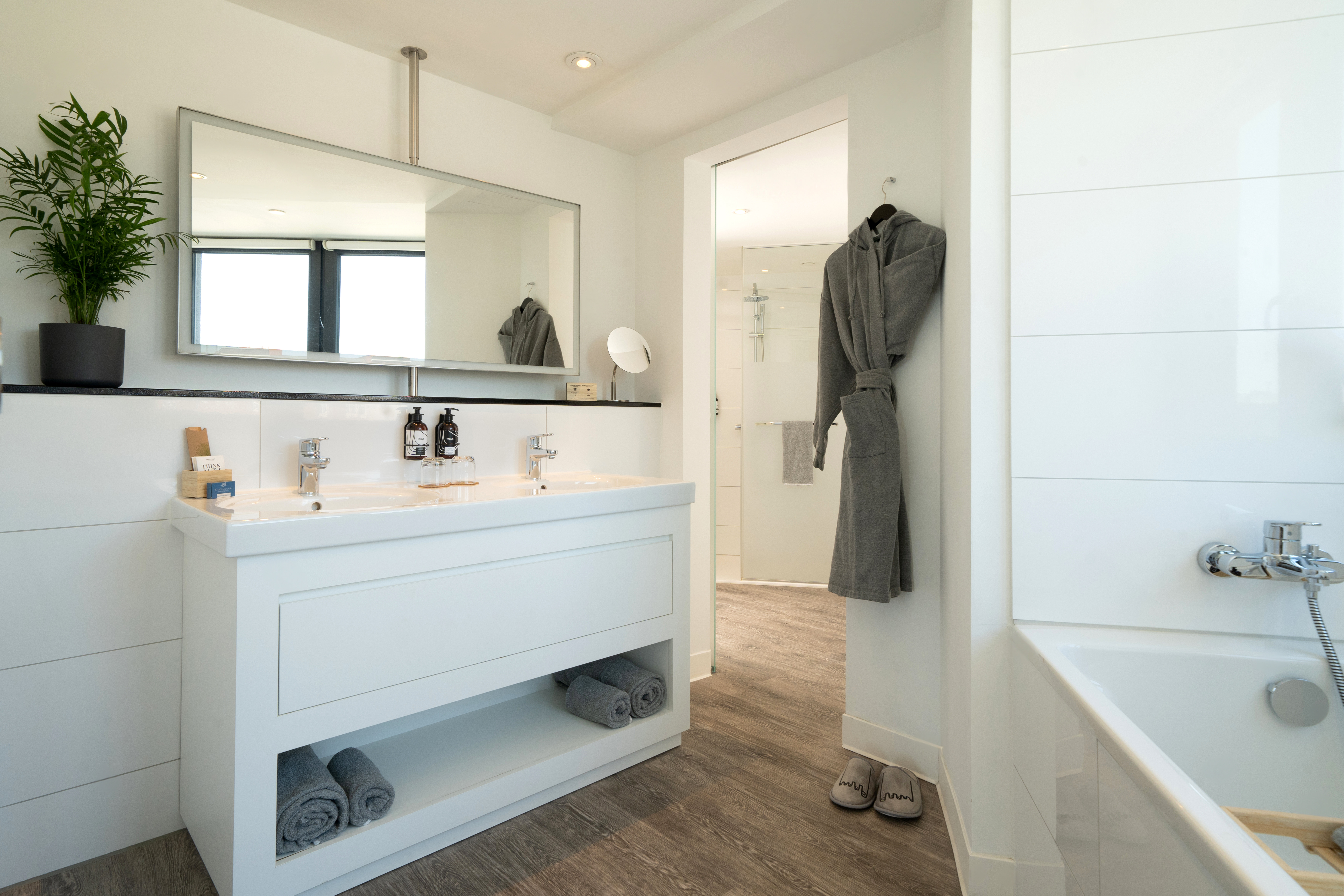 Sleek white bathroom with double vanity, large mirror, plant, and grey robe.