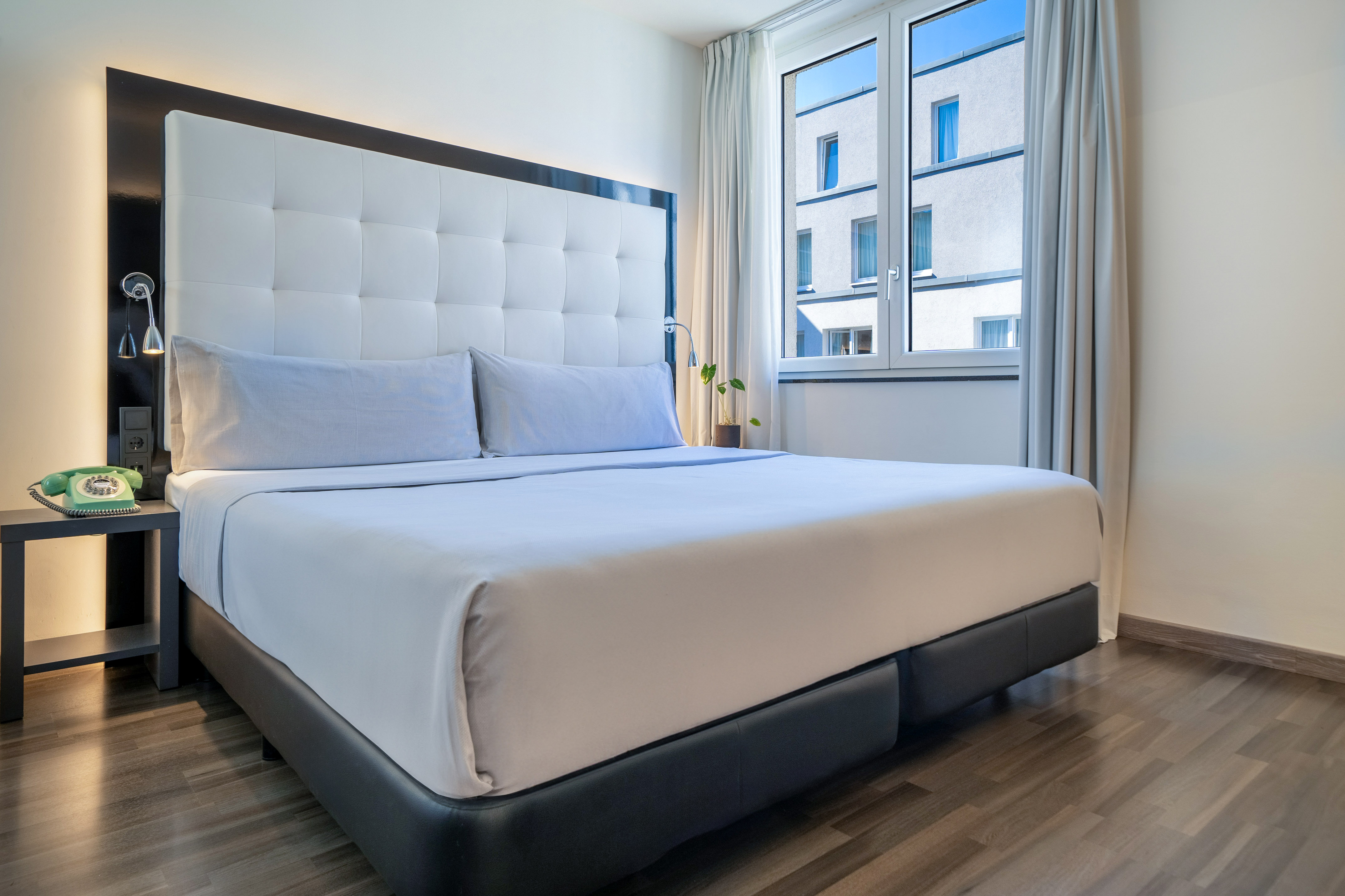 Modern hotel room with large bed, white headboard, blue sheets, and window.