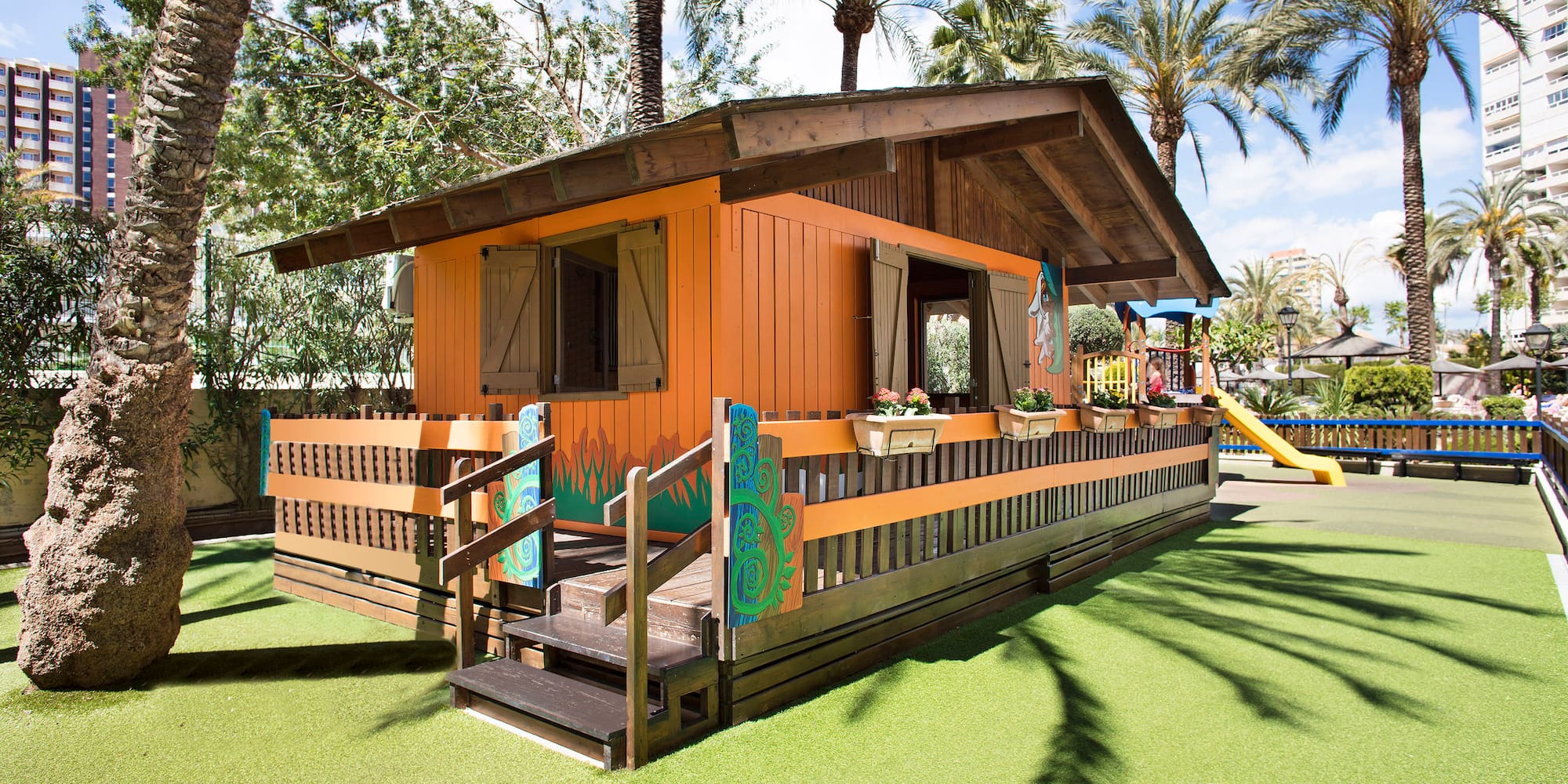 Casita de juegos infantil naranja en césped artificial con palmeras.