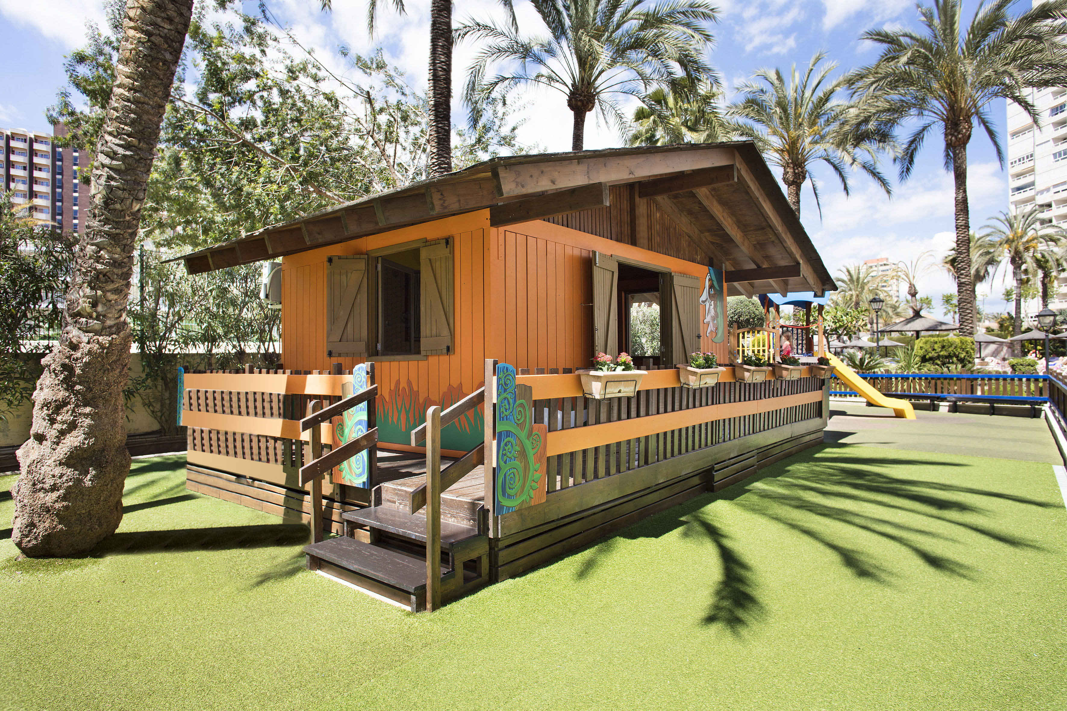 Casita de juegos infantil naranja en césped artificial con palmeras.