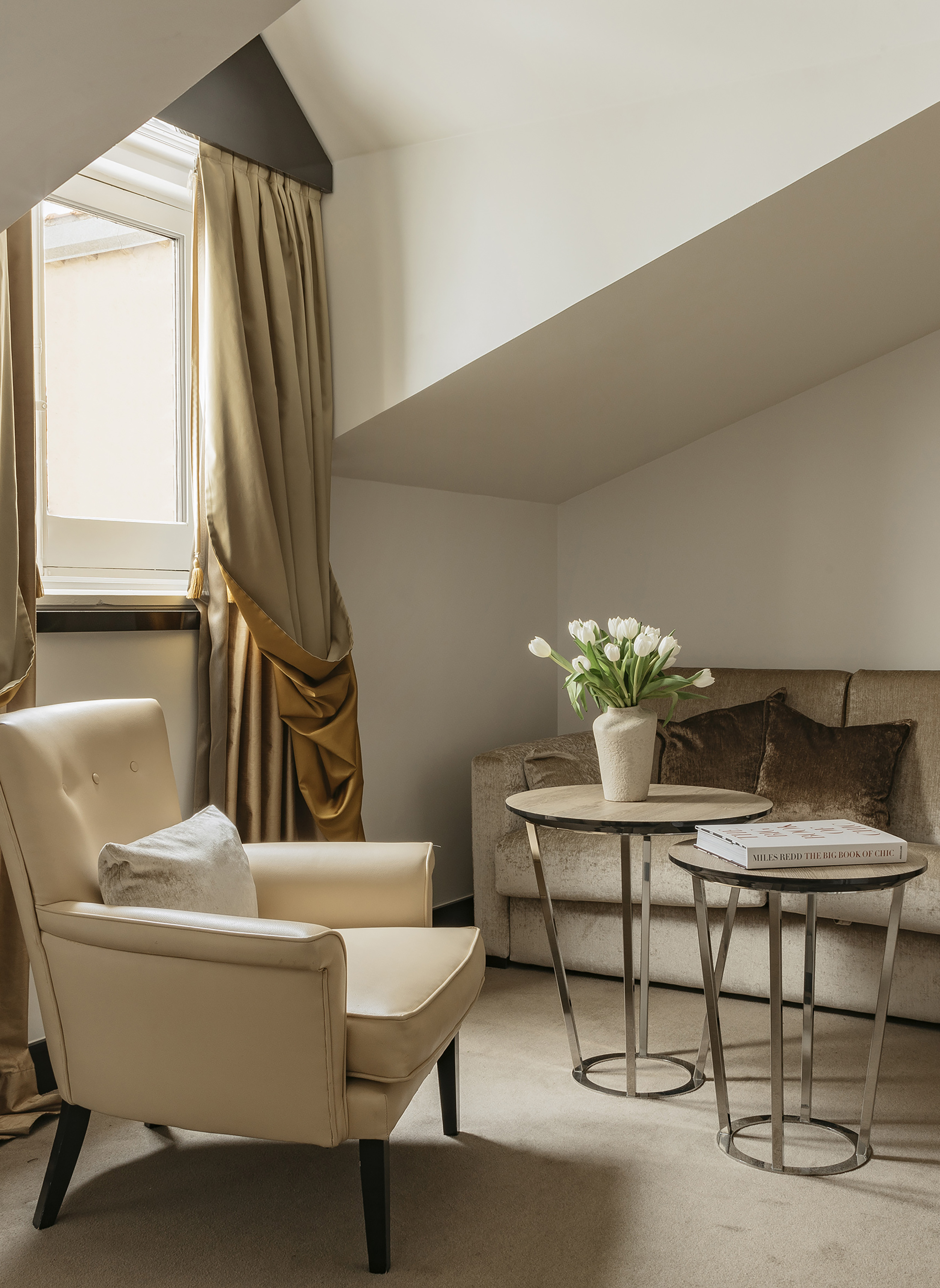 Attic living room with beige armchair, brown sofa, coffee tables, and white tulips.