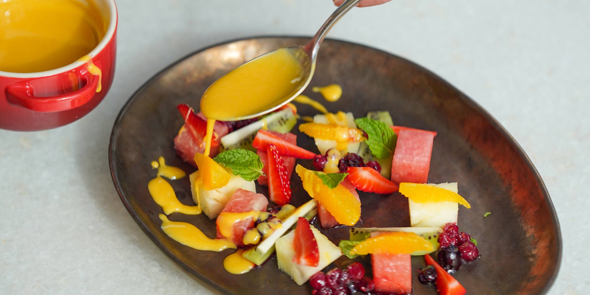 Hand pouring yellow dressing on colorful fresh fruit salad.