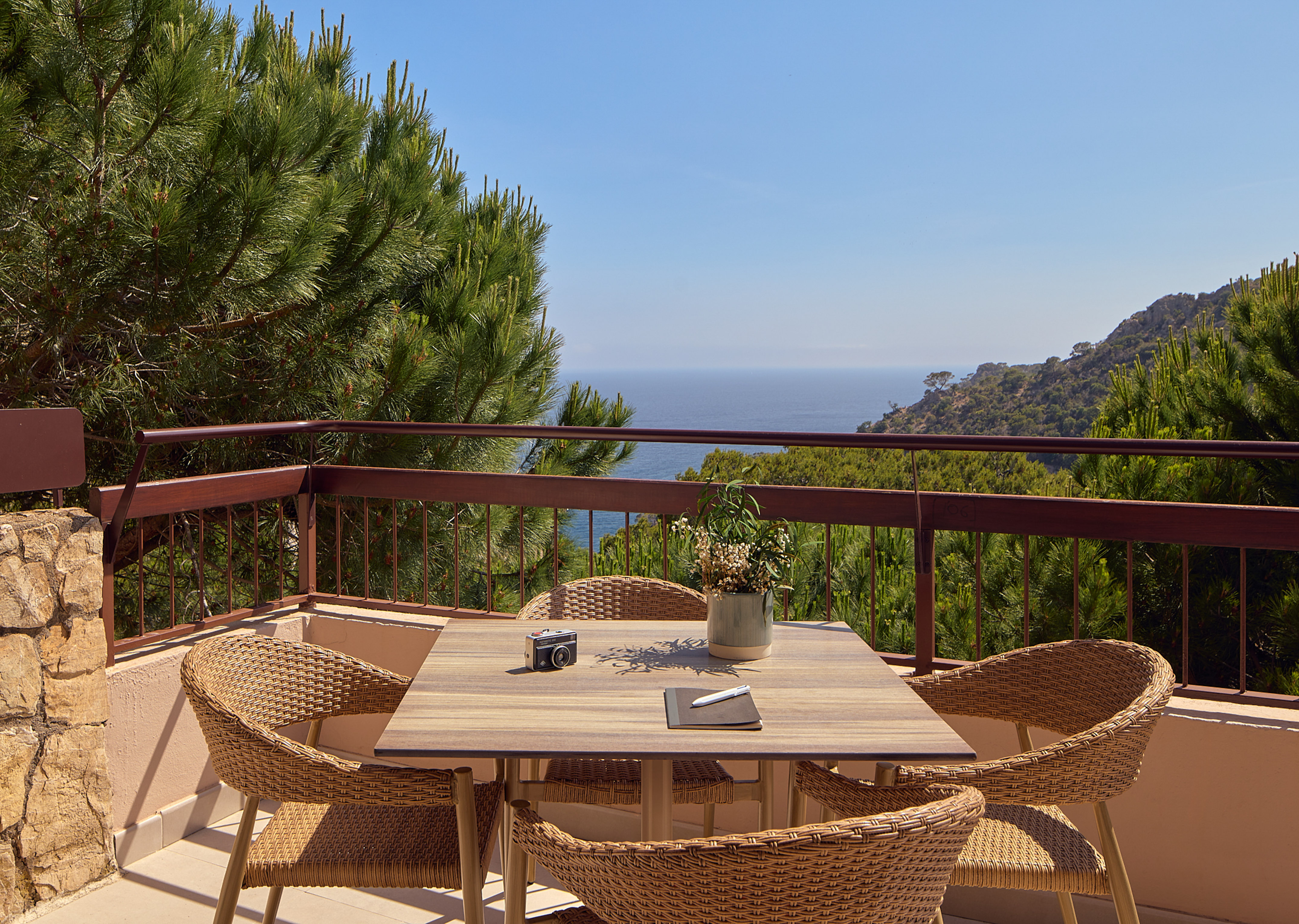 Balcony with outdoor table, wicker chairs, camera, overlooking coastal sea.