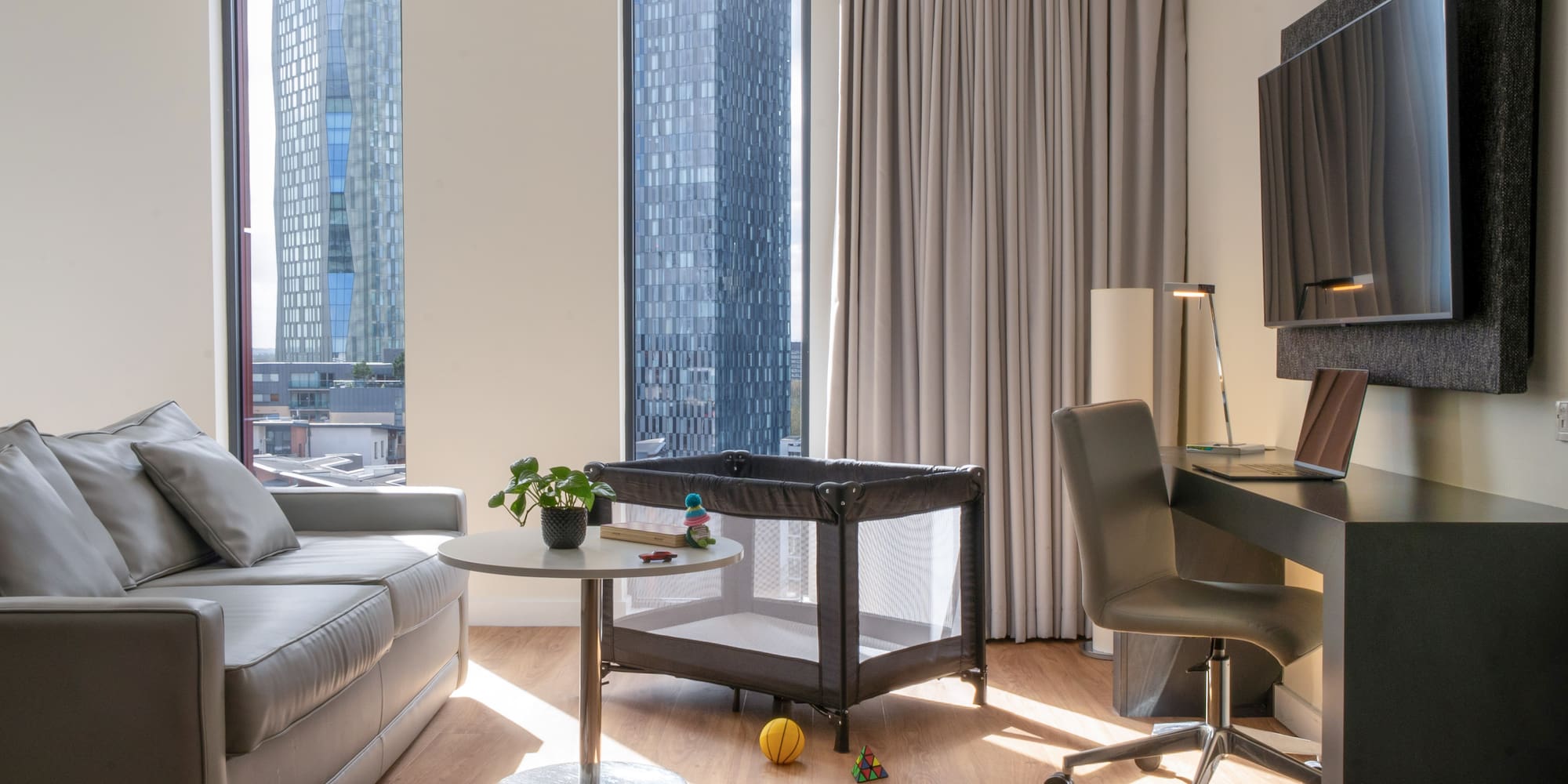 Modern hotel living area with sofa, desk, travel cot, and city skyline view.