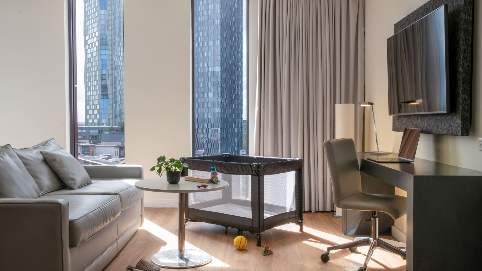 Modern hotel living area with sofa, desk, travel cot, and city skyline view.