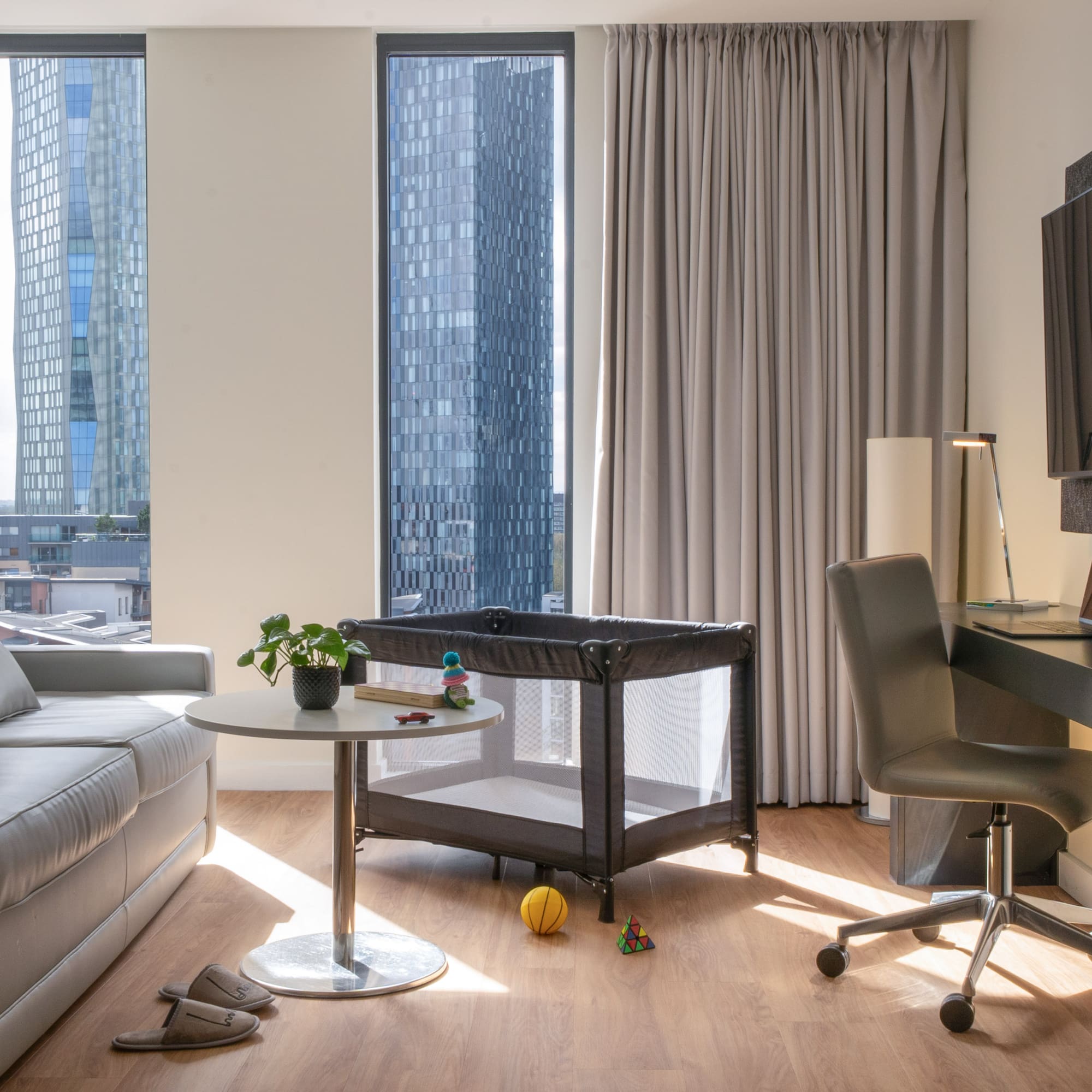 Modern hotel living area with sofa, desk, travel cot, and city skyline view.