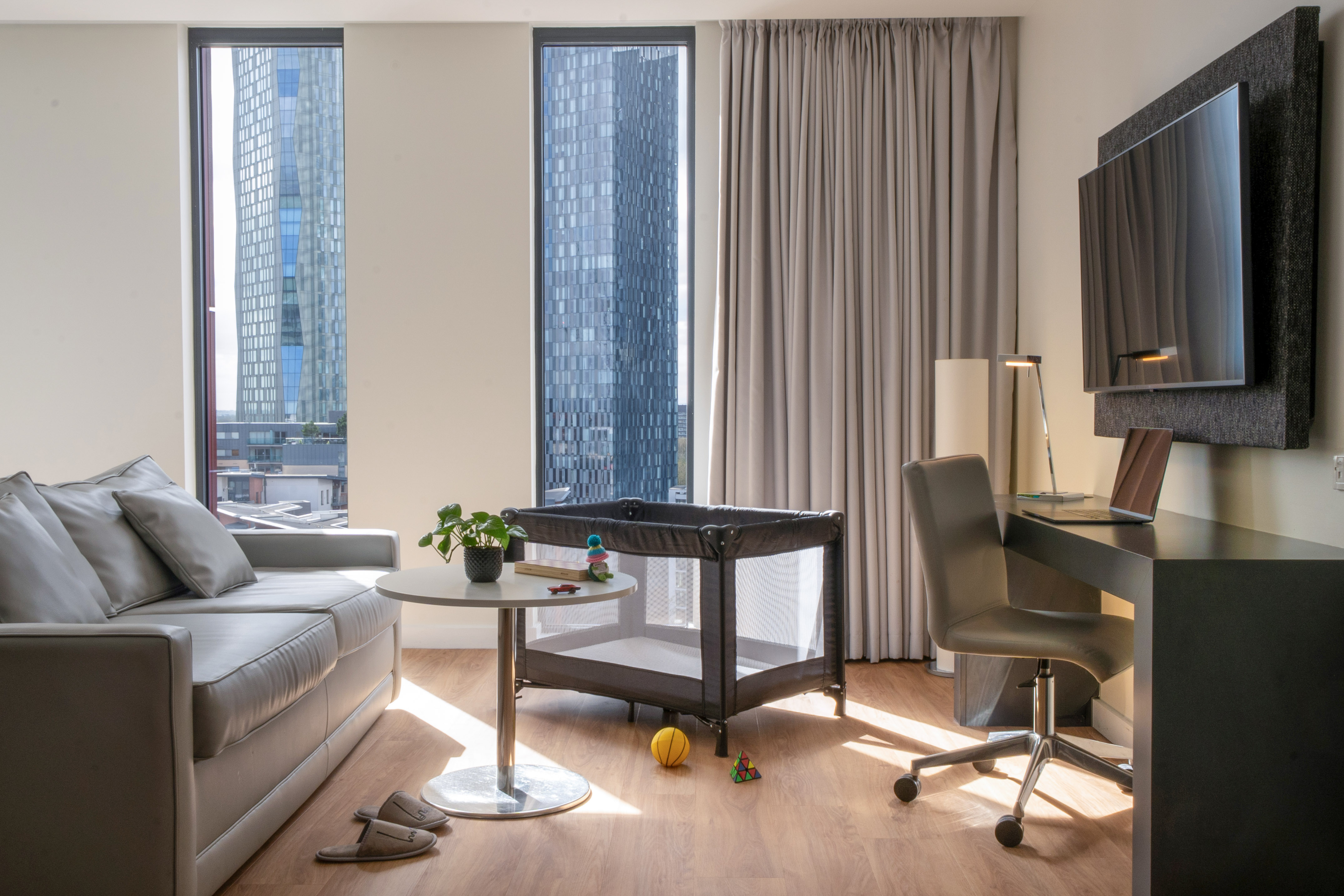 Modern hotel living area with sofa, desk, travel cot, and city skyline view.