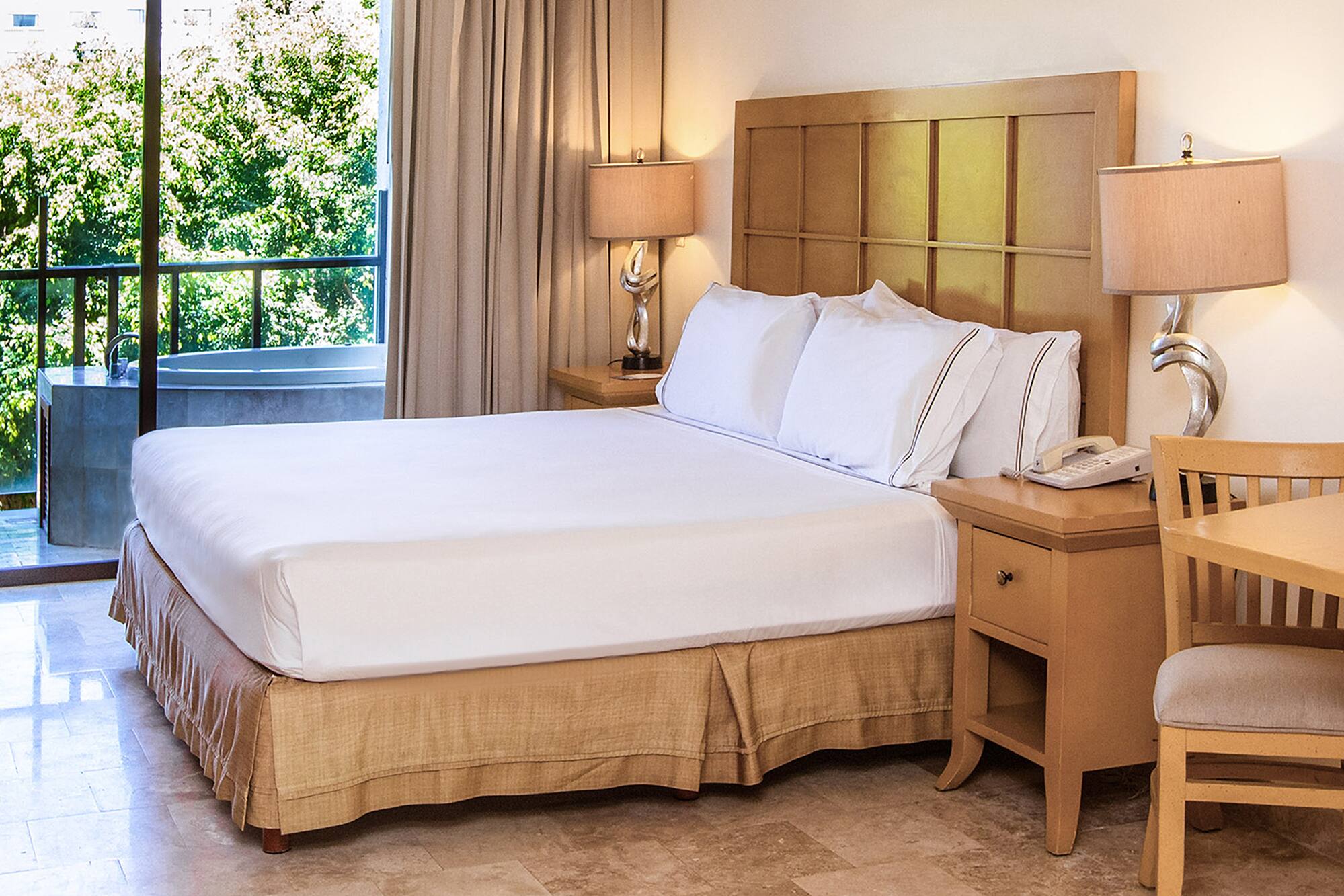 Hotel bedroom with king bed, nightstands, balcony spa tub, and green view.
