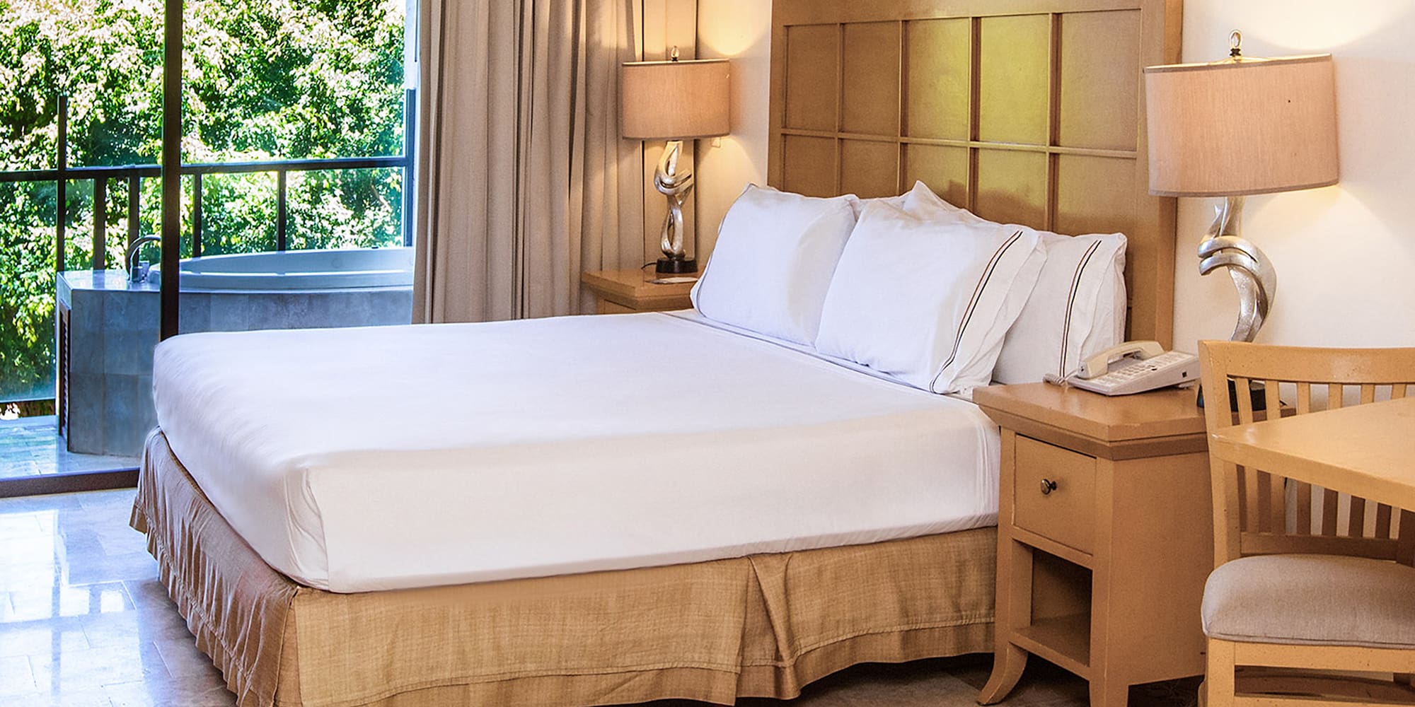 Hotel bedroom with king bed, nightstands, balcony spa tub, and green view.