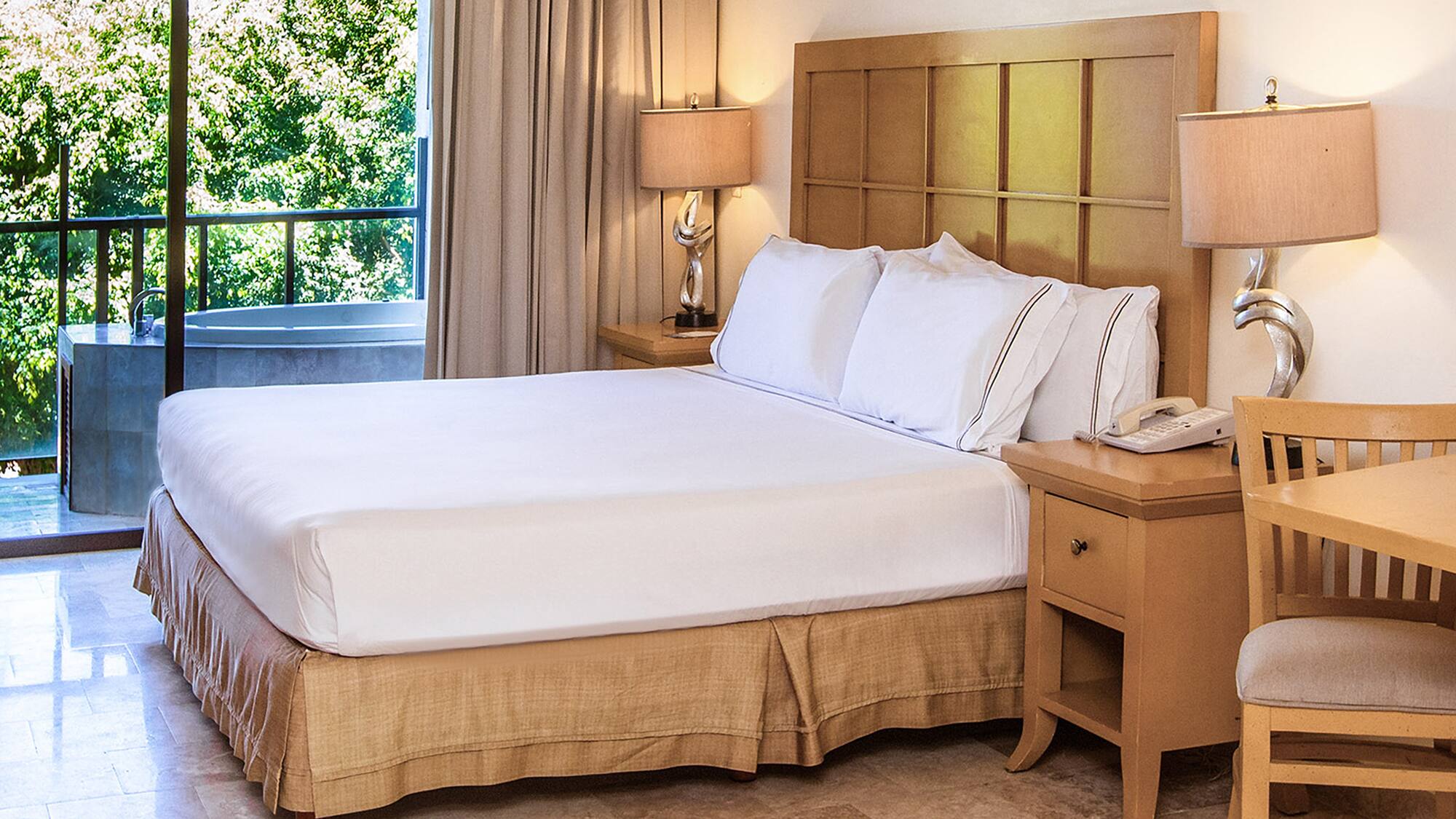 Hotel bedroom with king bed, nightstands, balcony spa tub, and green view.