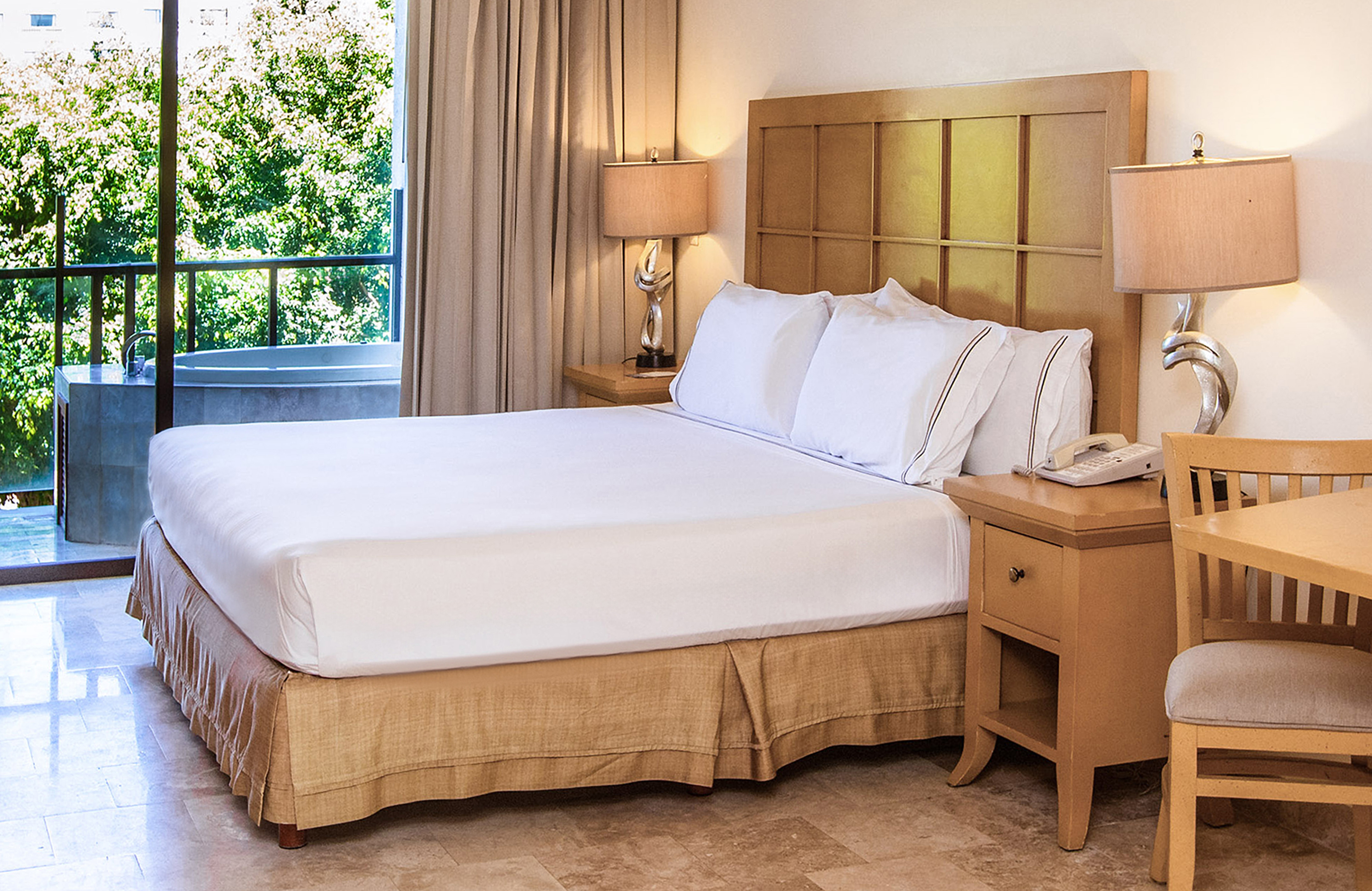 Hotel bedroom with king bed, nightstands, balcony spa tub, and green view.