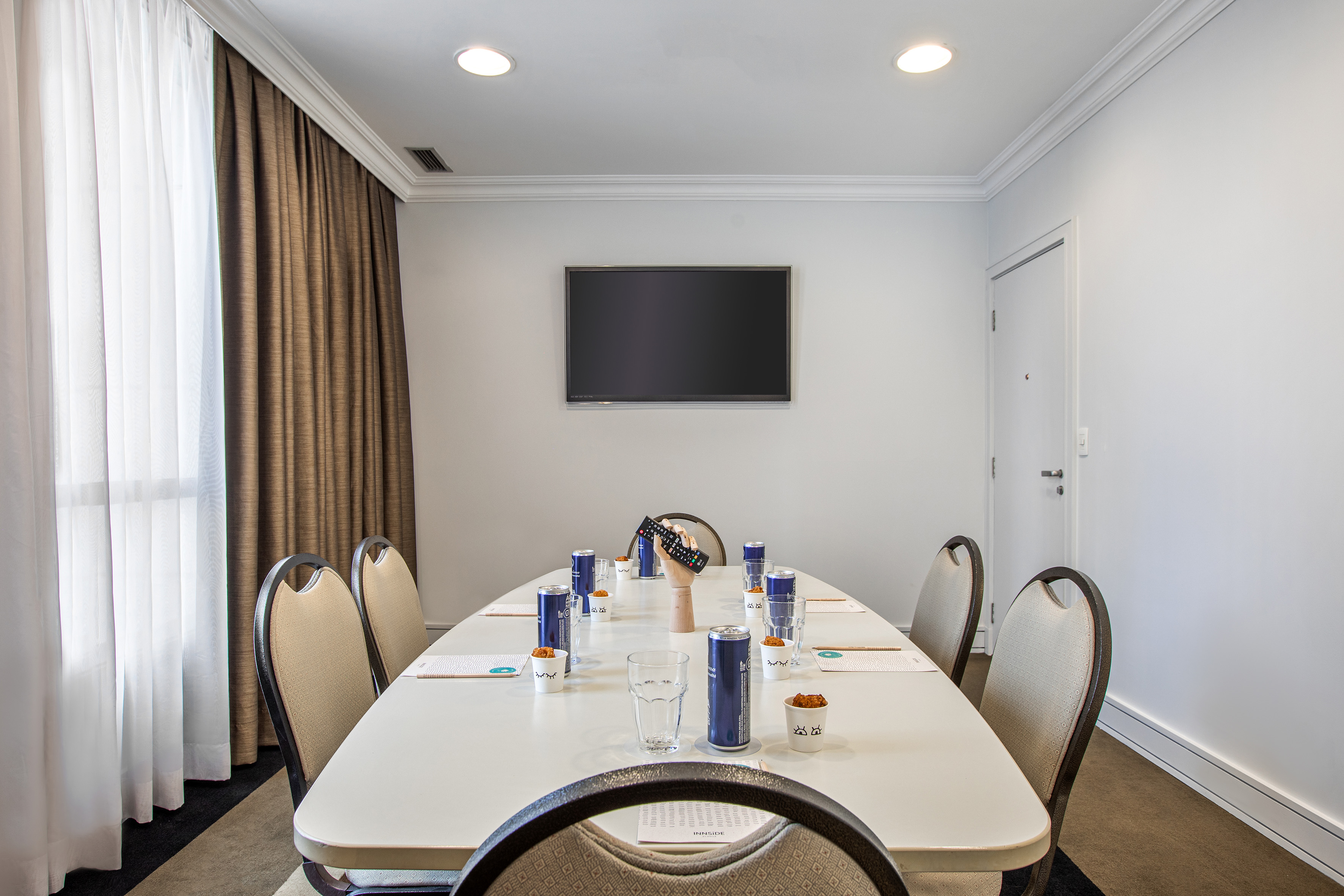Modern meeting room with conference table, chairs, wall TV, and refreshments.