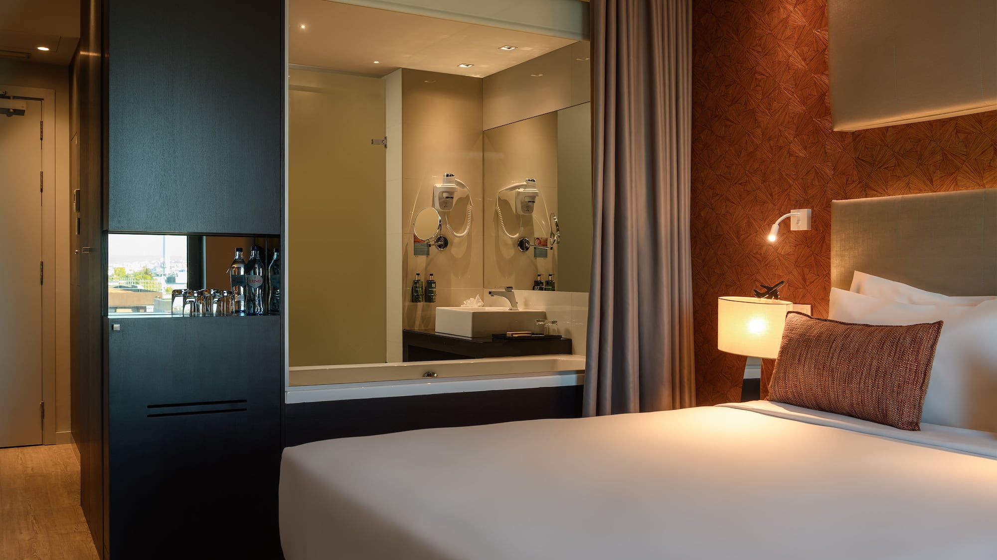 Modern hotel room with bed, open-concept bathroom, and refreshment cabinet.
