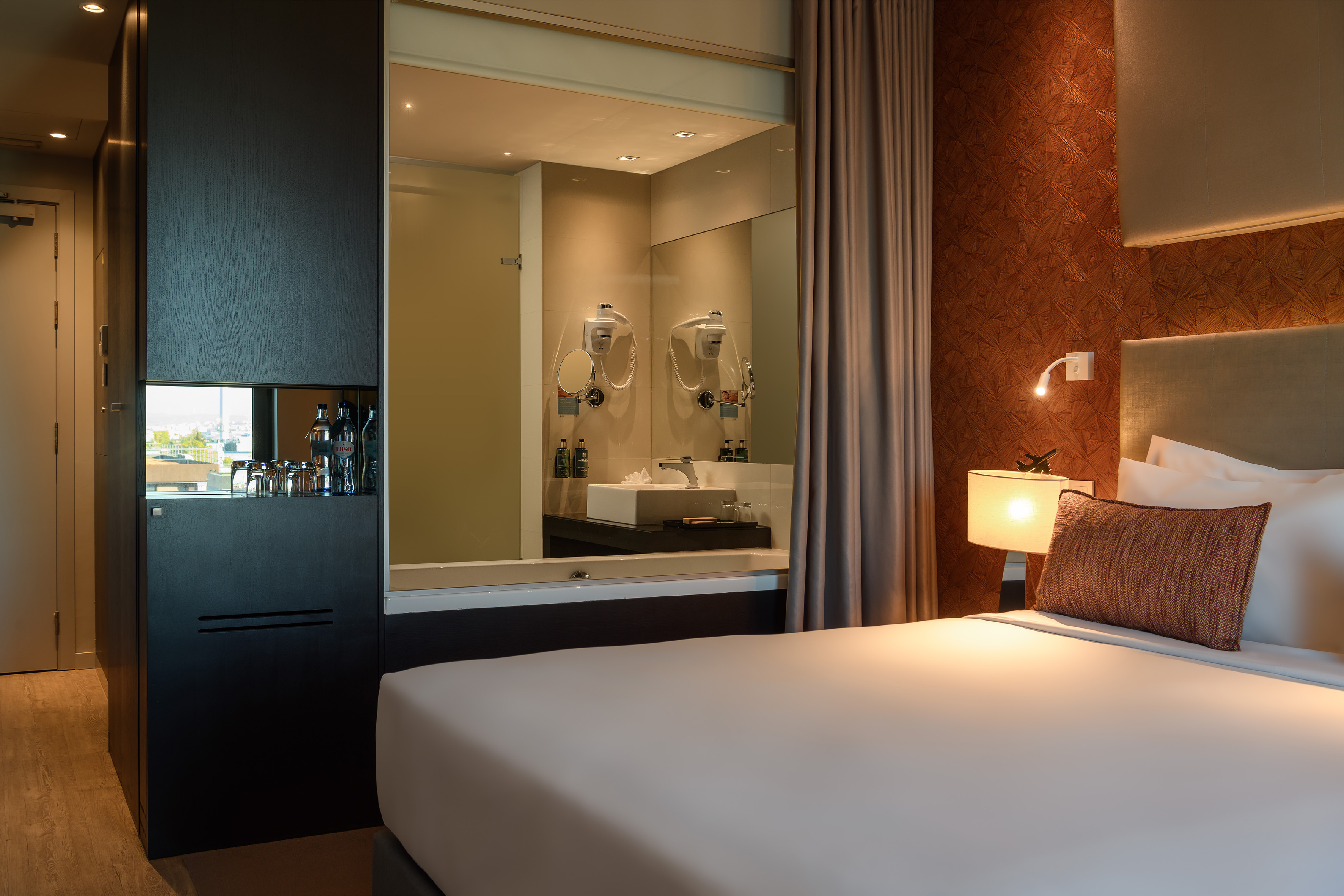 Modern hotel room with bed, open-concept bathroom, and refreshment cabinet.