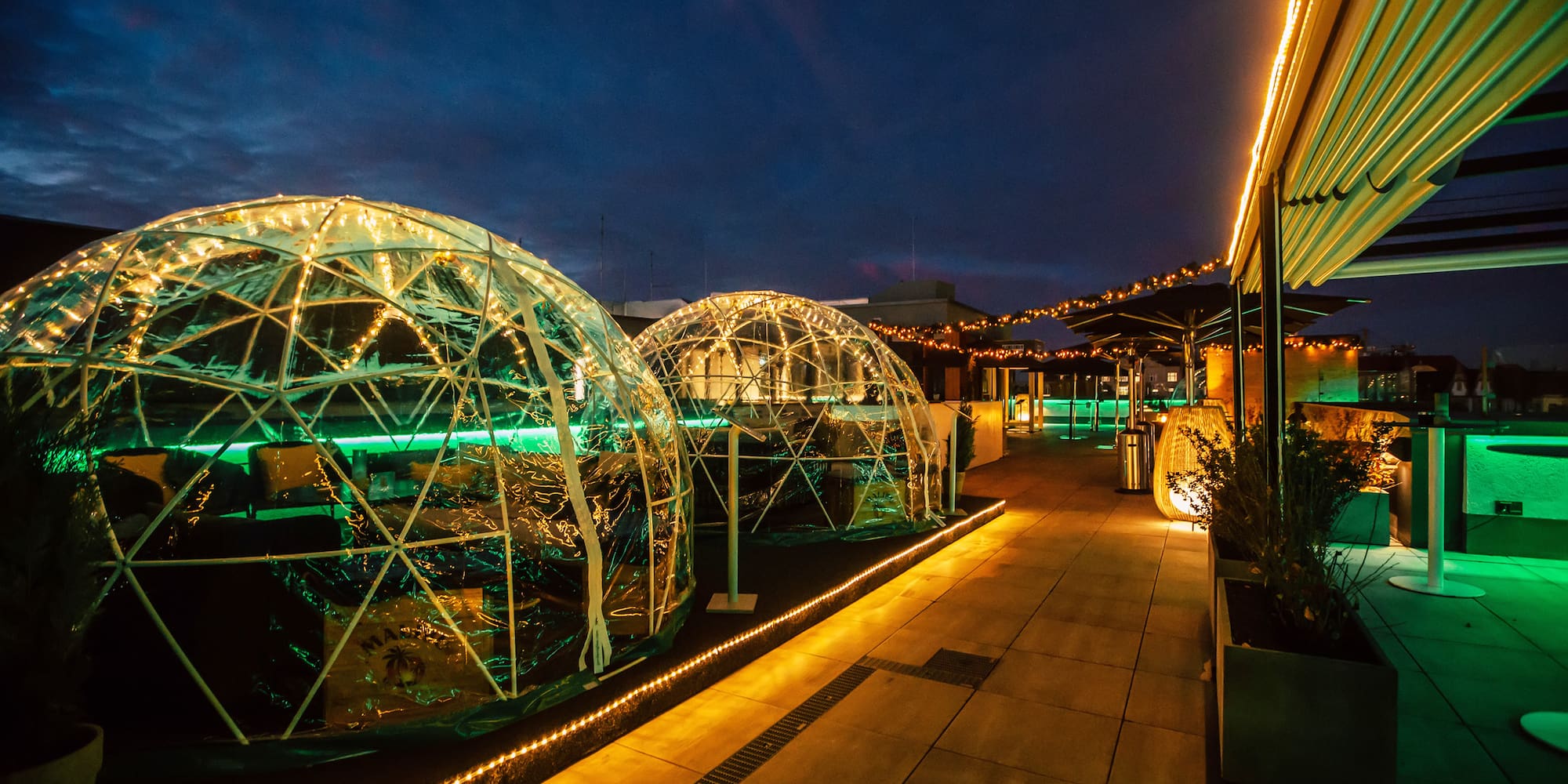 Rooftop bar at dusk with glowing geodesic domes, ambient green lighting.