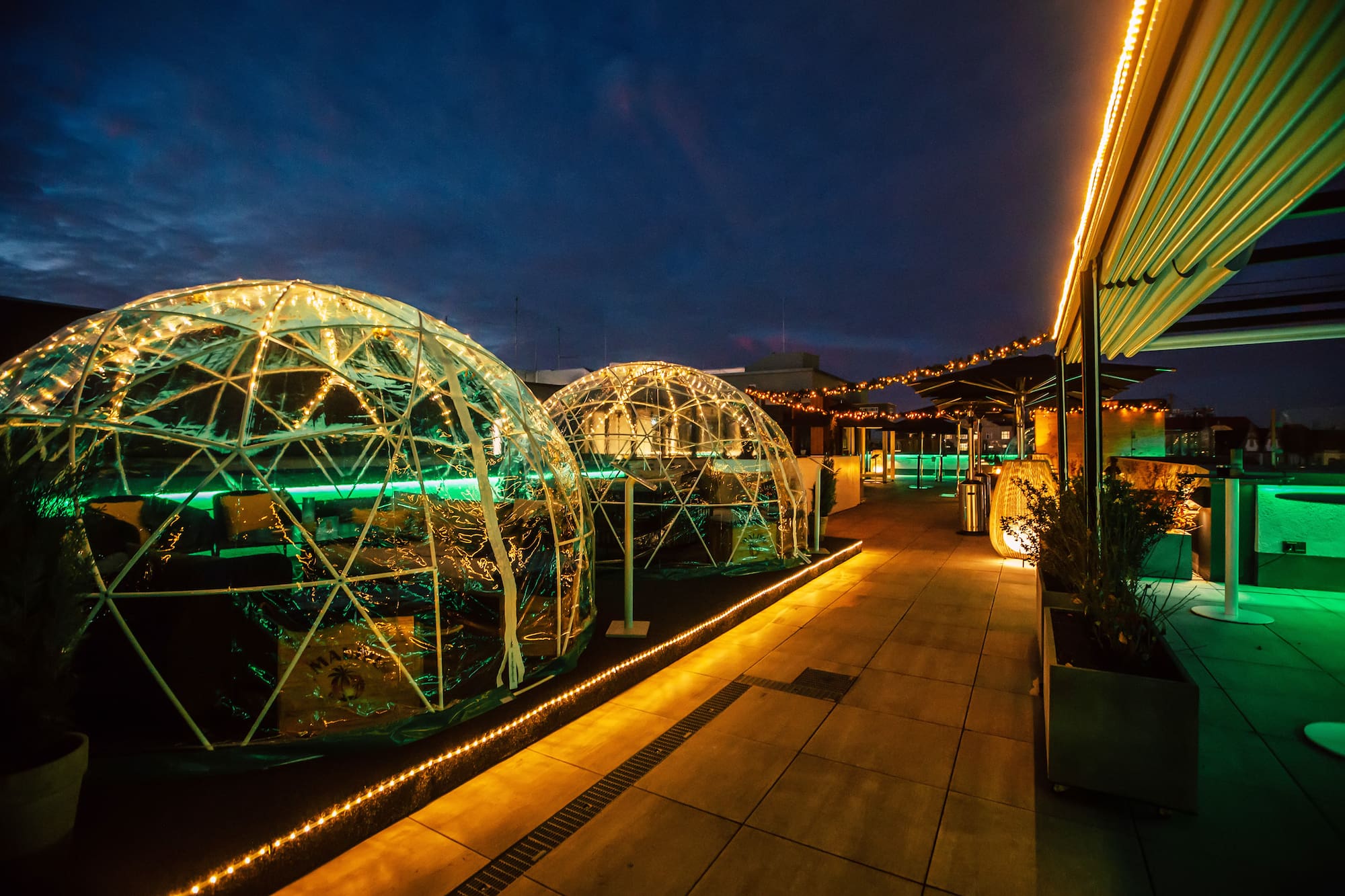 Rooftop bar at dusk with glowing geodesic domes, ambient green lighting.