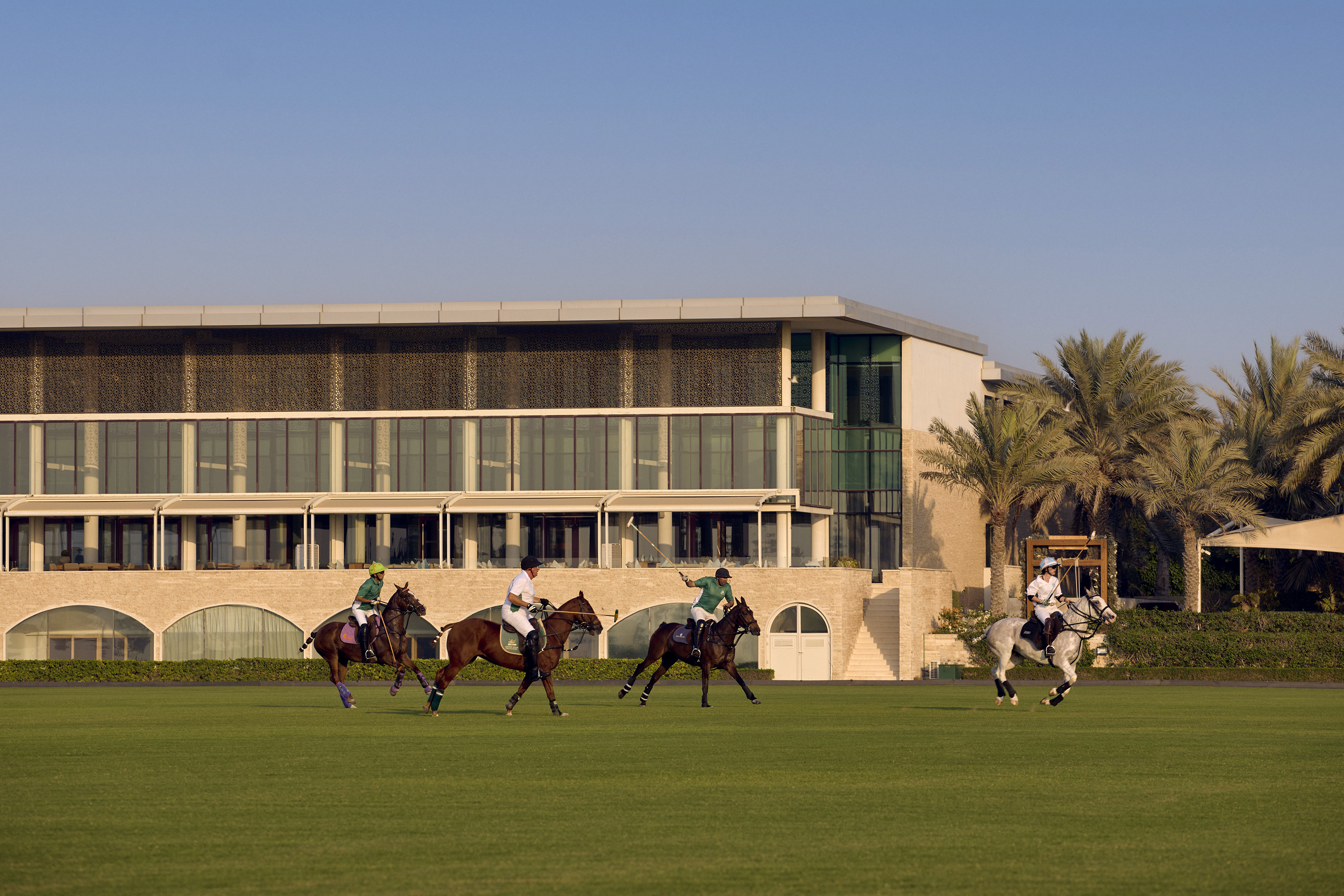 Four polo players on horseback on a green field with a modern building and palm trees.