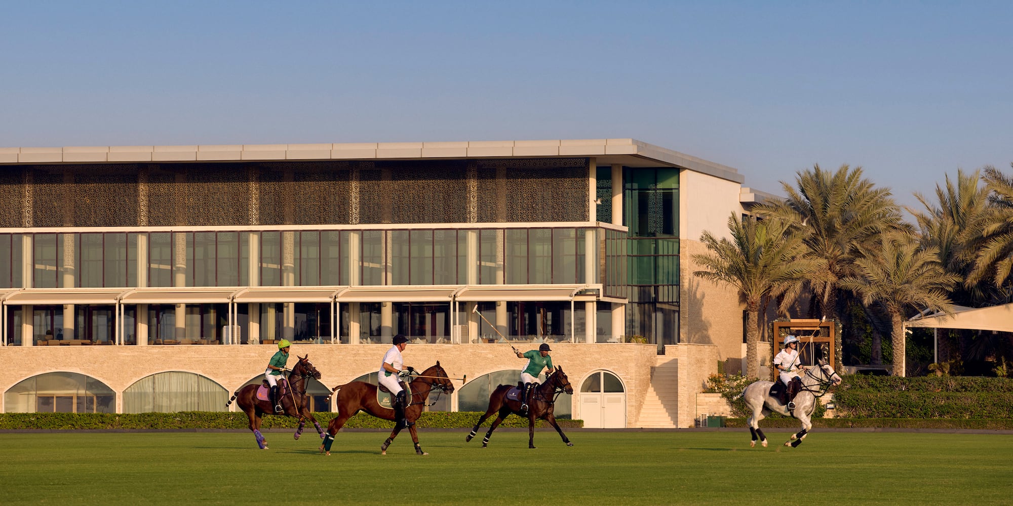 Four polo players on horseback on a green field with a modern building and palm trees.