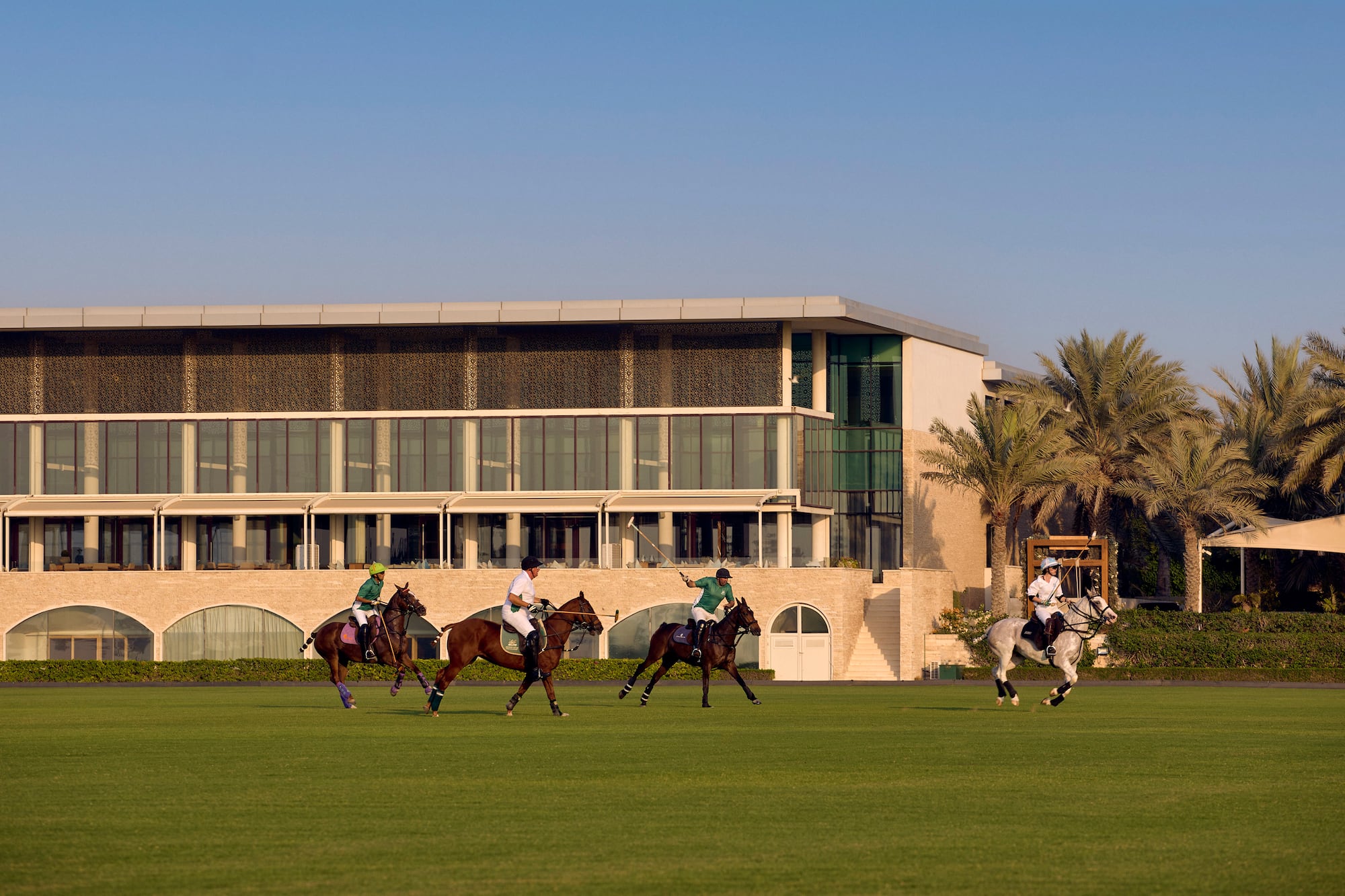Four polo players on horseback on a green field with a modern building and palm trees.