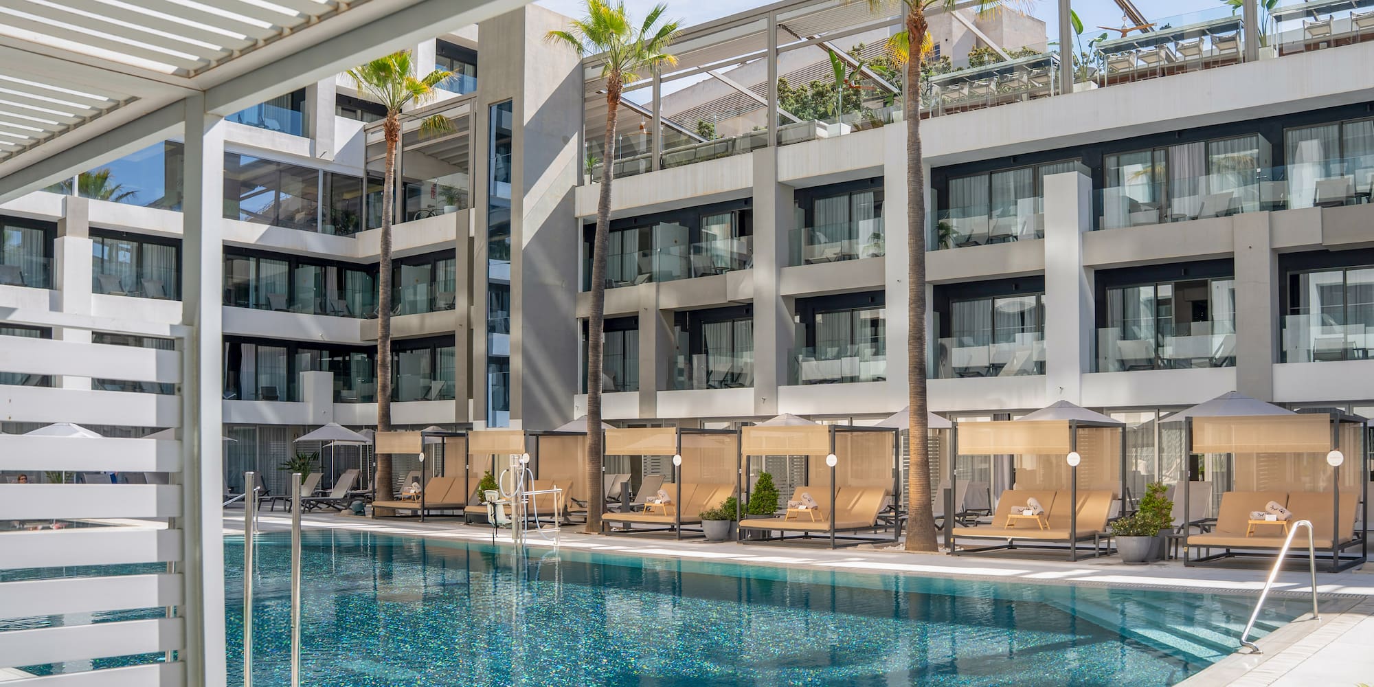 Luxury resort pool with modern hotel building, palm trees, and shaded cabanas.
