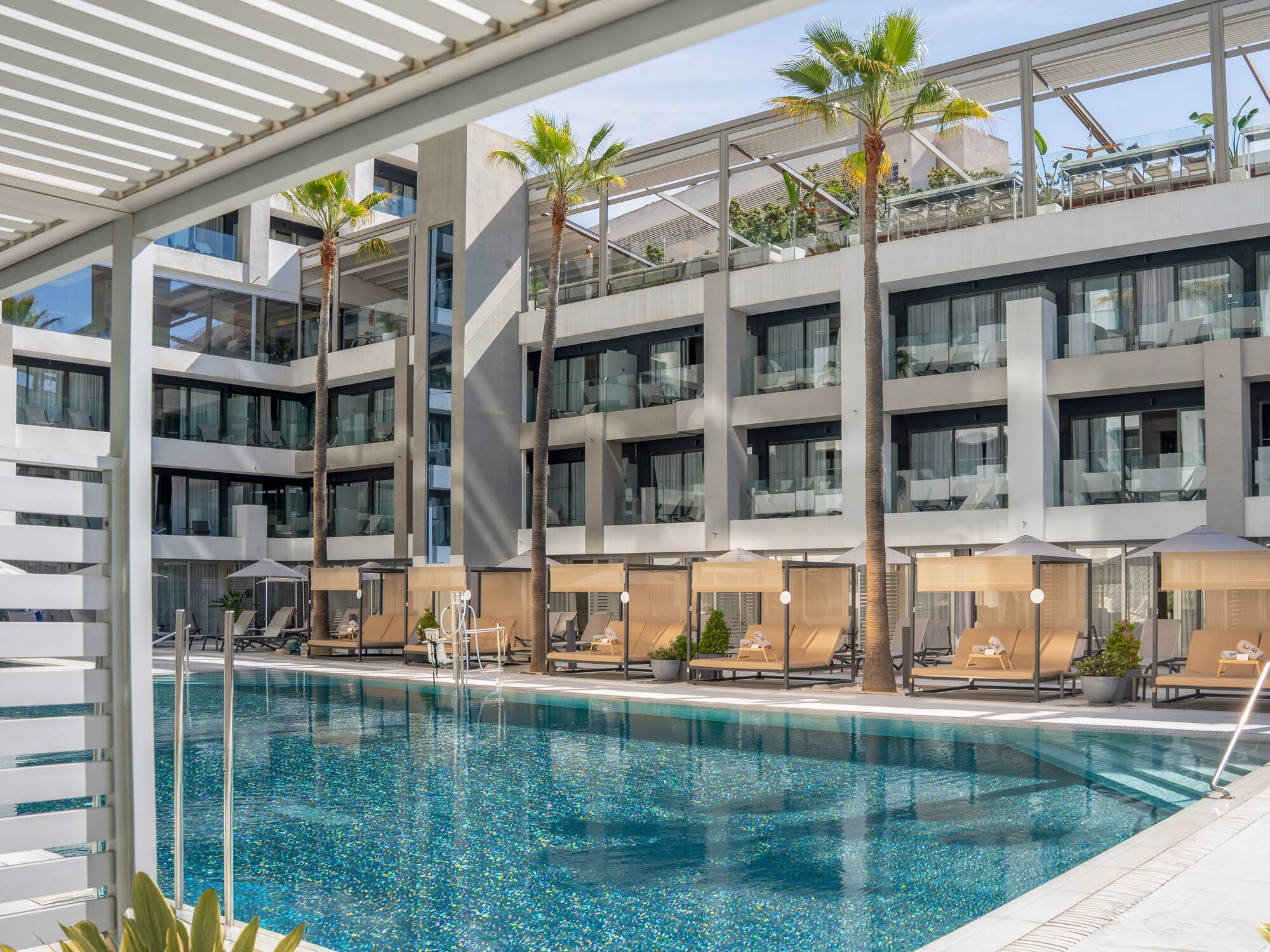 Luxury resort pool with modern hotel building, palm trees, and shaded cabanas.