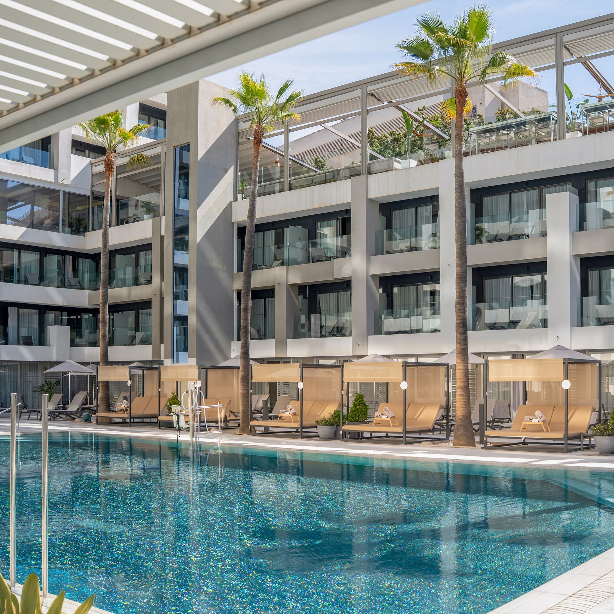 Sparkling blue hotel pool with modern architecture, palm trees, and poolside daybeds.