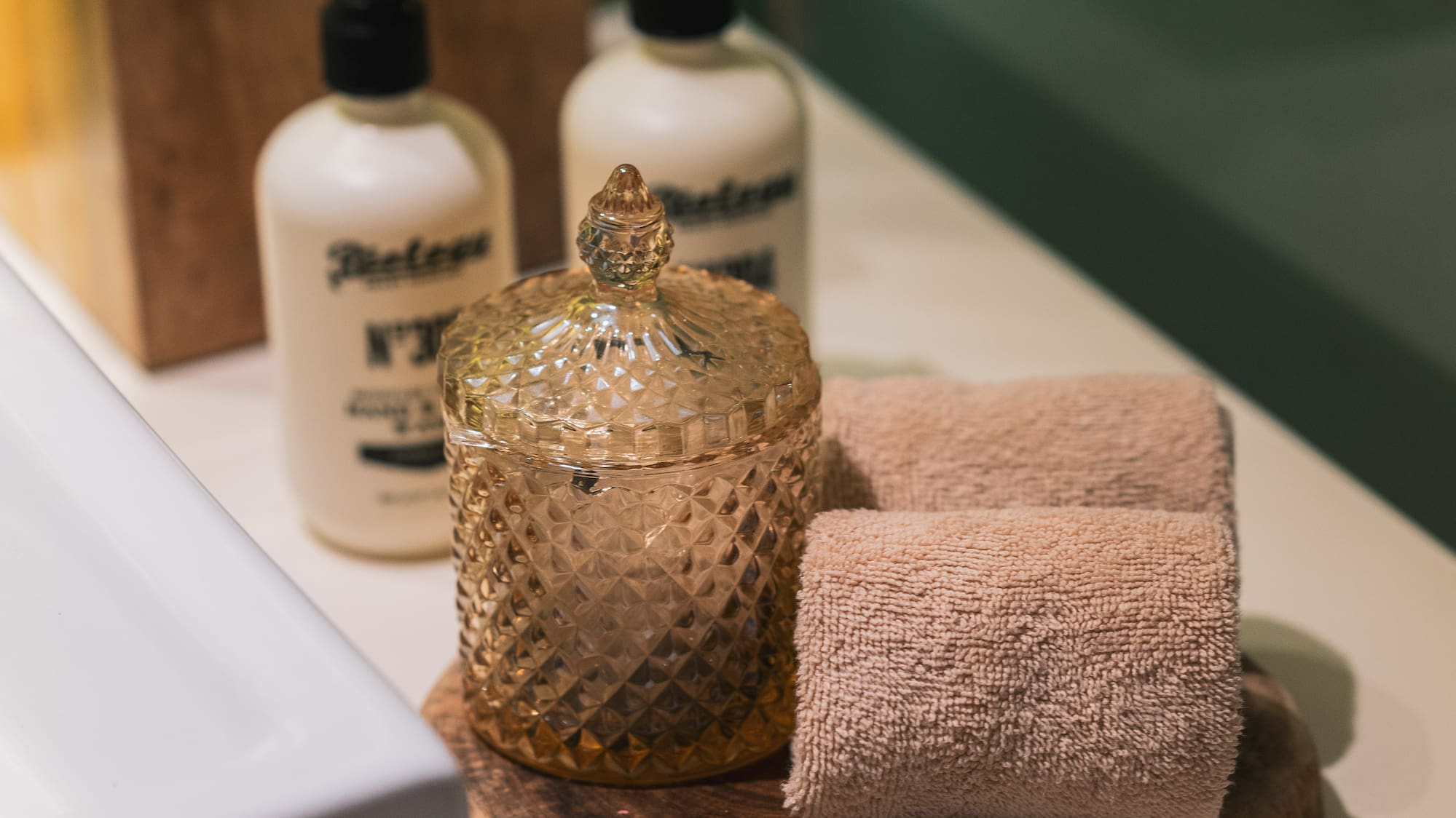 Bathroom vanity with white soap bottles, wooden tissue box, amber jar, and rolled towels.
