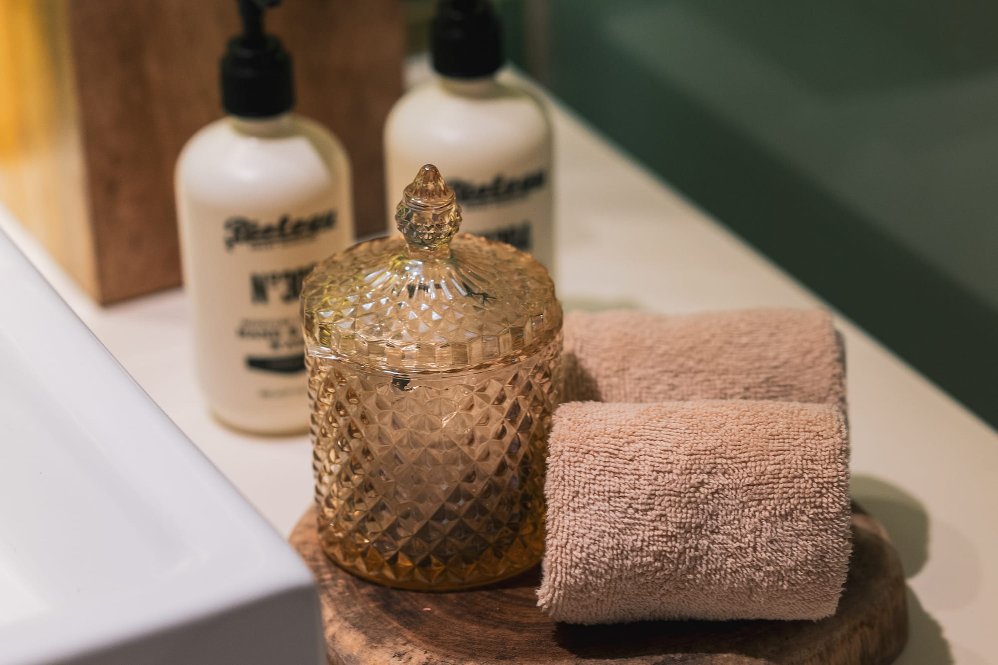 Bathroom vanity with white soap bottles, wooden tissue box, amber jar, and rolled towels.