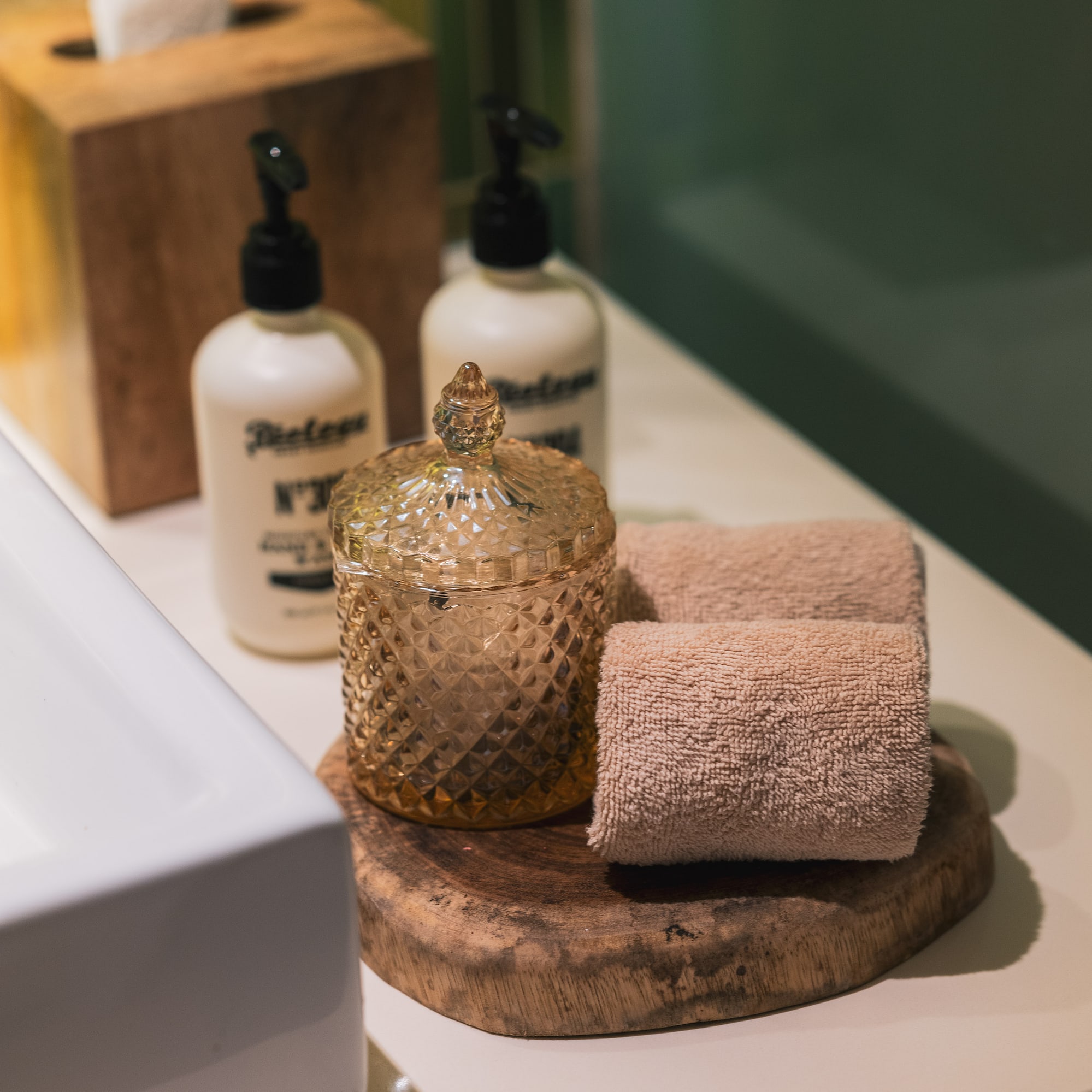 Bathroom vanity with white soap bottles, wooden tissue box, amber jar, and rolled towels.