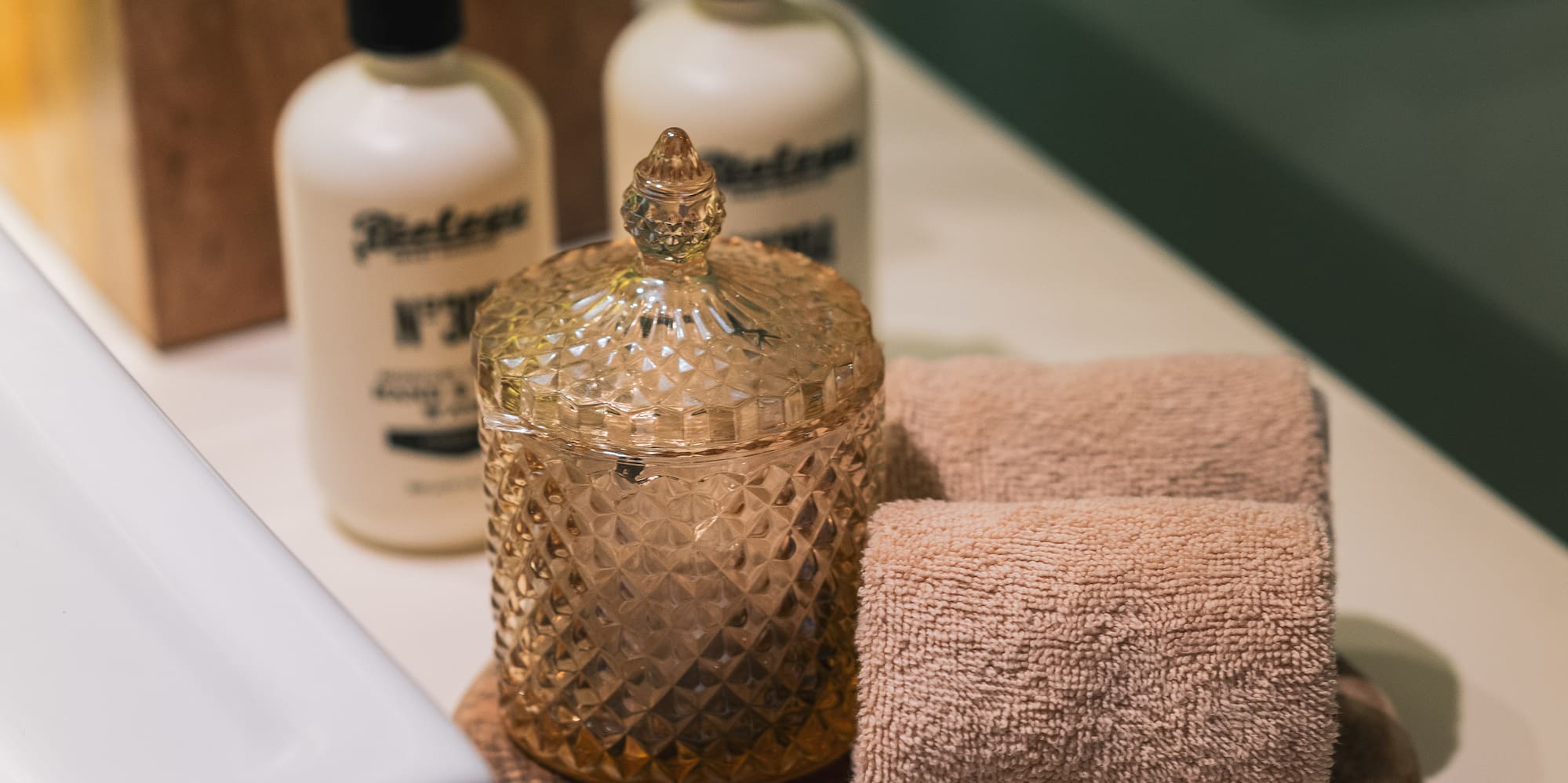 Bathroom vanity with white soap bottles, wooden tissue box, amber jar, and rolled towels.