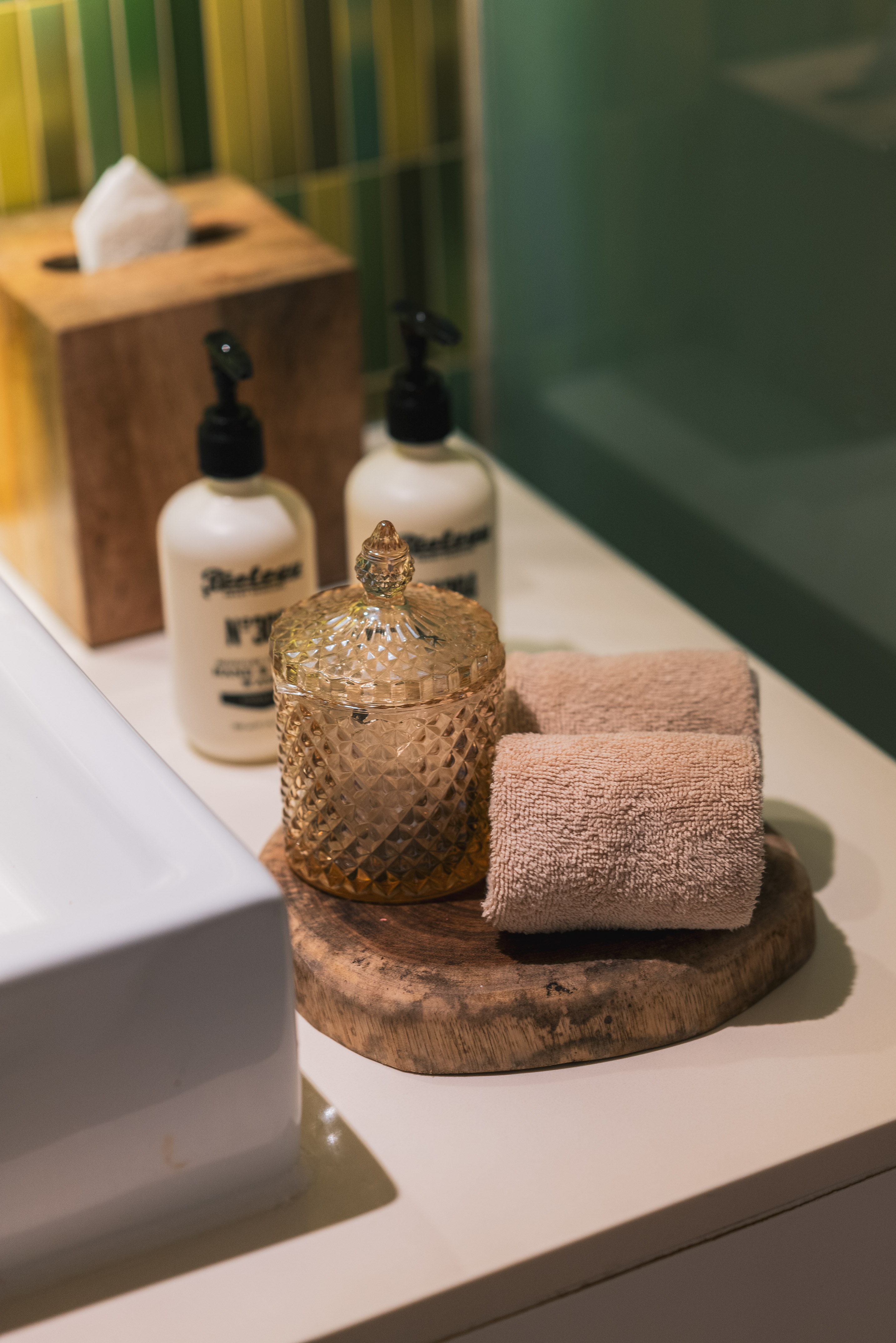 Bathroom vanity with white soap bottles, wooden tissue box, amber jar, and rolled towels.