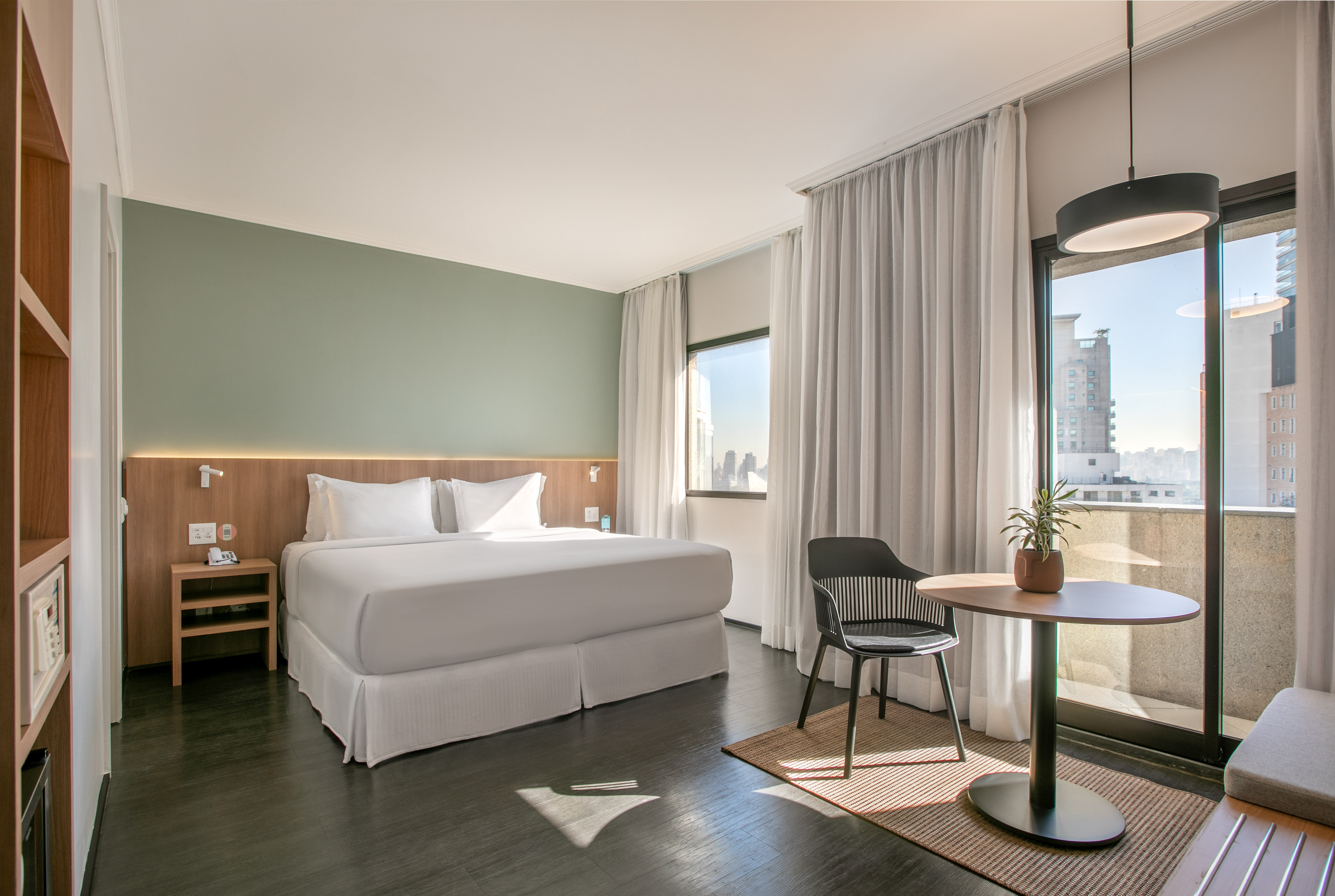 Modern hotel room with bed, green accent wall, city view window, seating area.