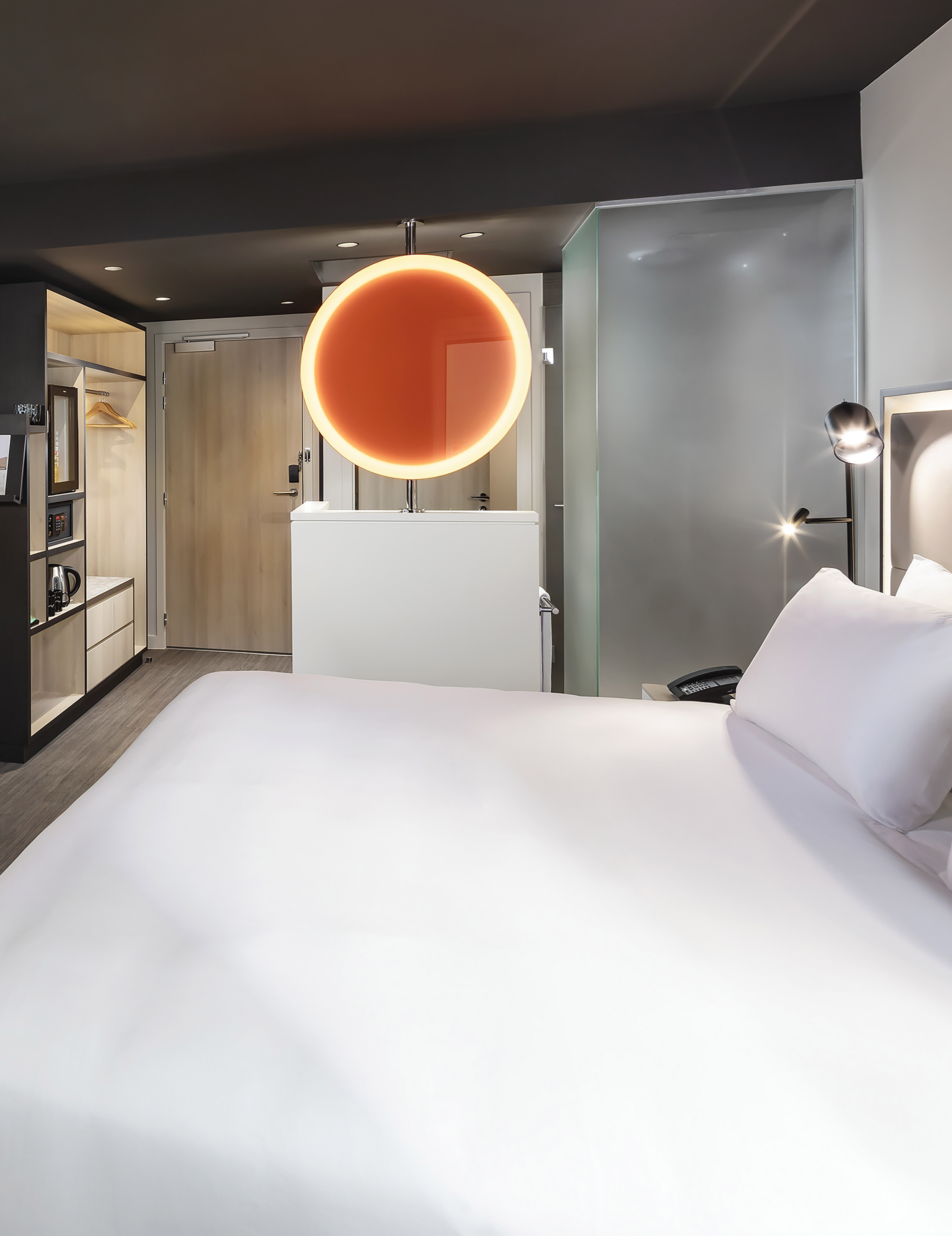 Modern hotel room with white bed, orange circular light, wardrobe, and frosted glass.